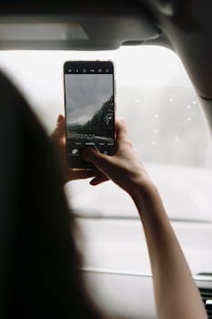 Person taking photo of scenic drive through car window, featuring trees.