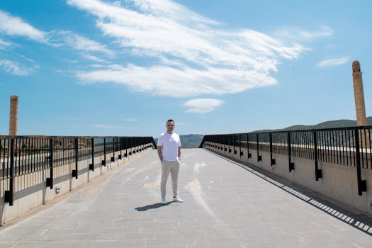 Man in white outfit standing on a bridge with clear blue sky and distant hills.
