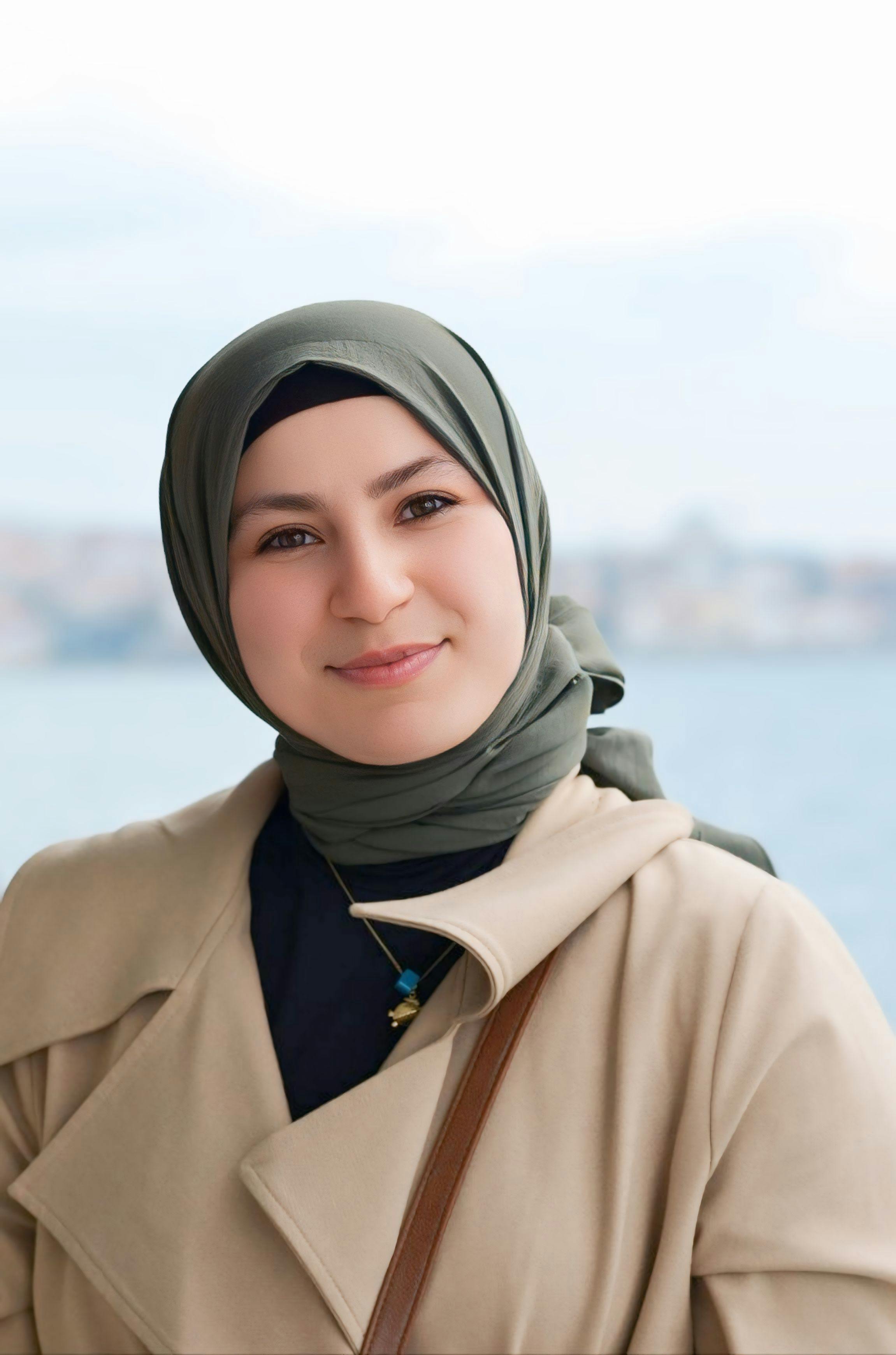 Portrait of a smiling woman in a green hijab outdoors with a cityscape backdrop.