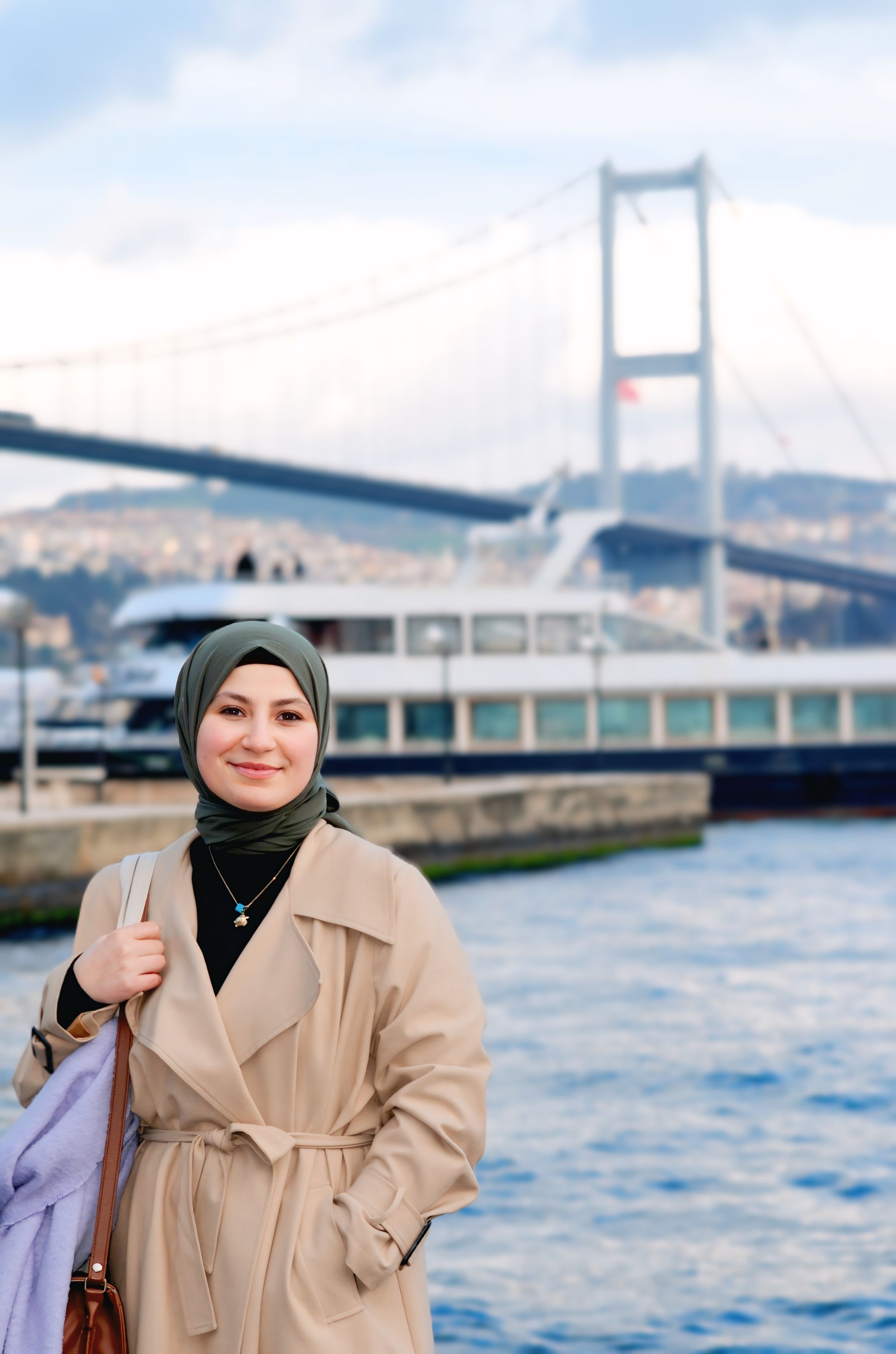 A smiling woman in hijab stands by the Bosphorus with the Bridge in Istanbul, Turkey.