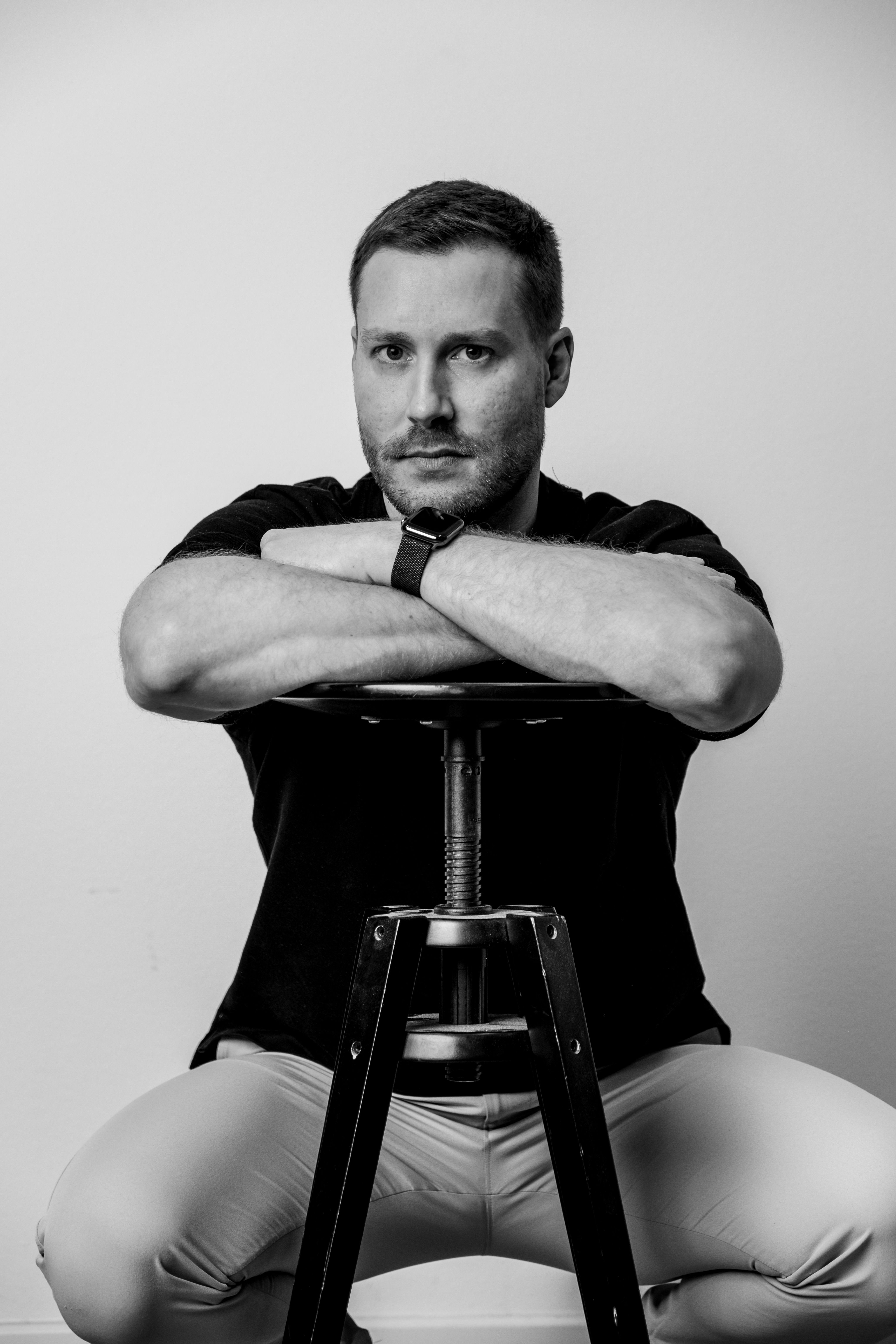 Monochrome portrait of a man seated, resting on a stool, looking directly at the camera.