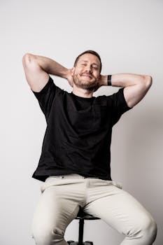 A bearded man casually sits on a stool in a black shirt, smiling and relaxed.