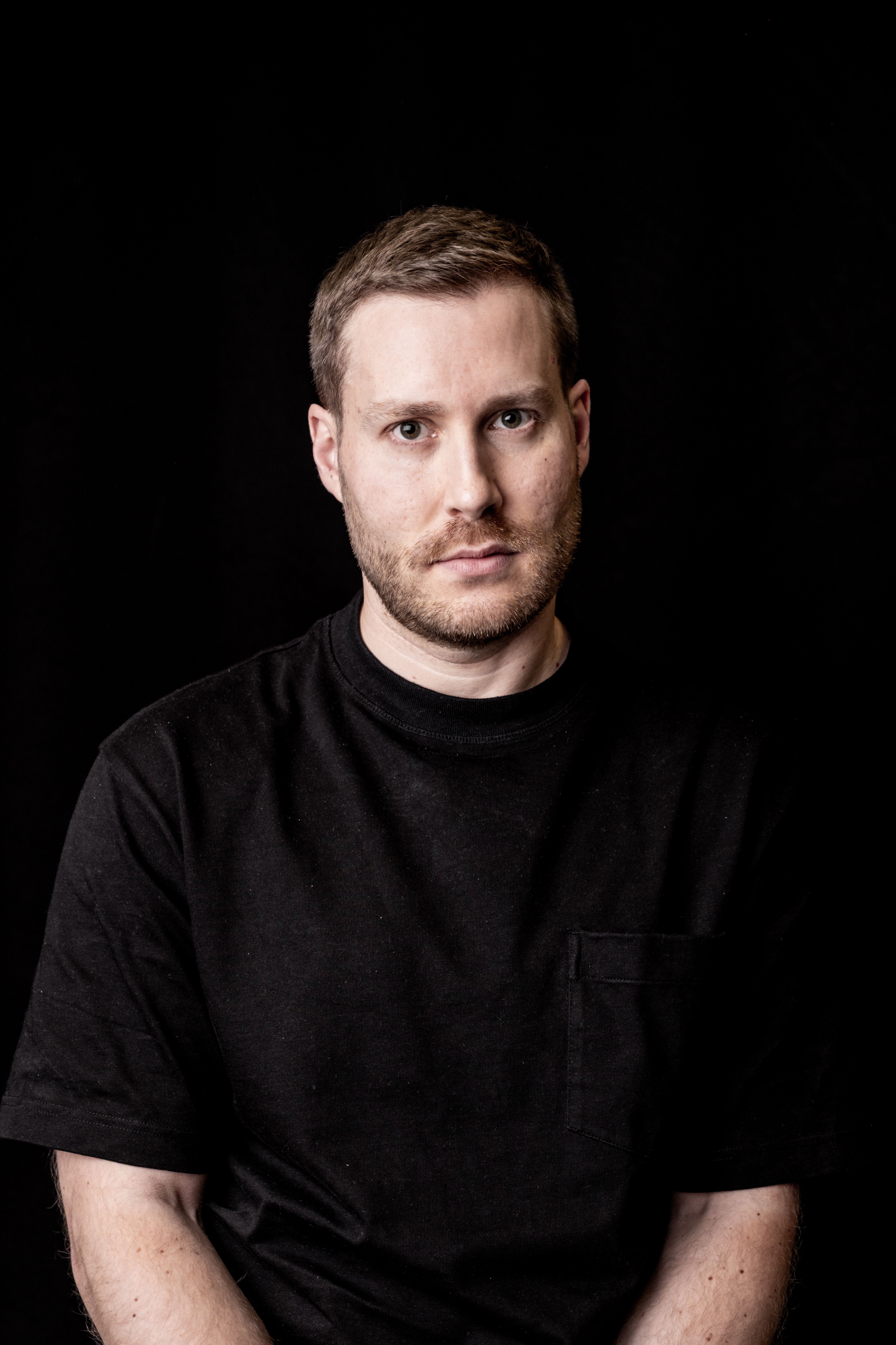 Studio portrait of a man wearing a black t-shirt, set against a dark background.