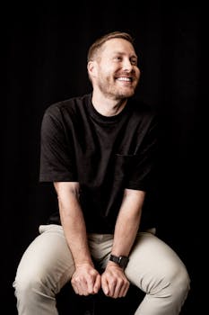 Caucasian man smiling against a dark backdrop wearing casual clothes.