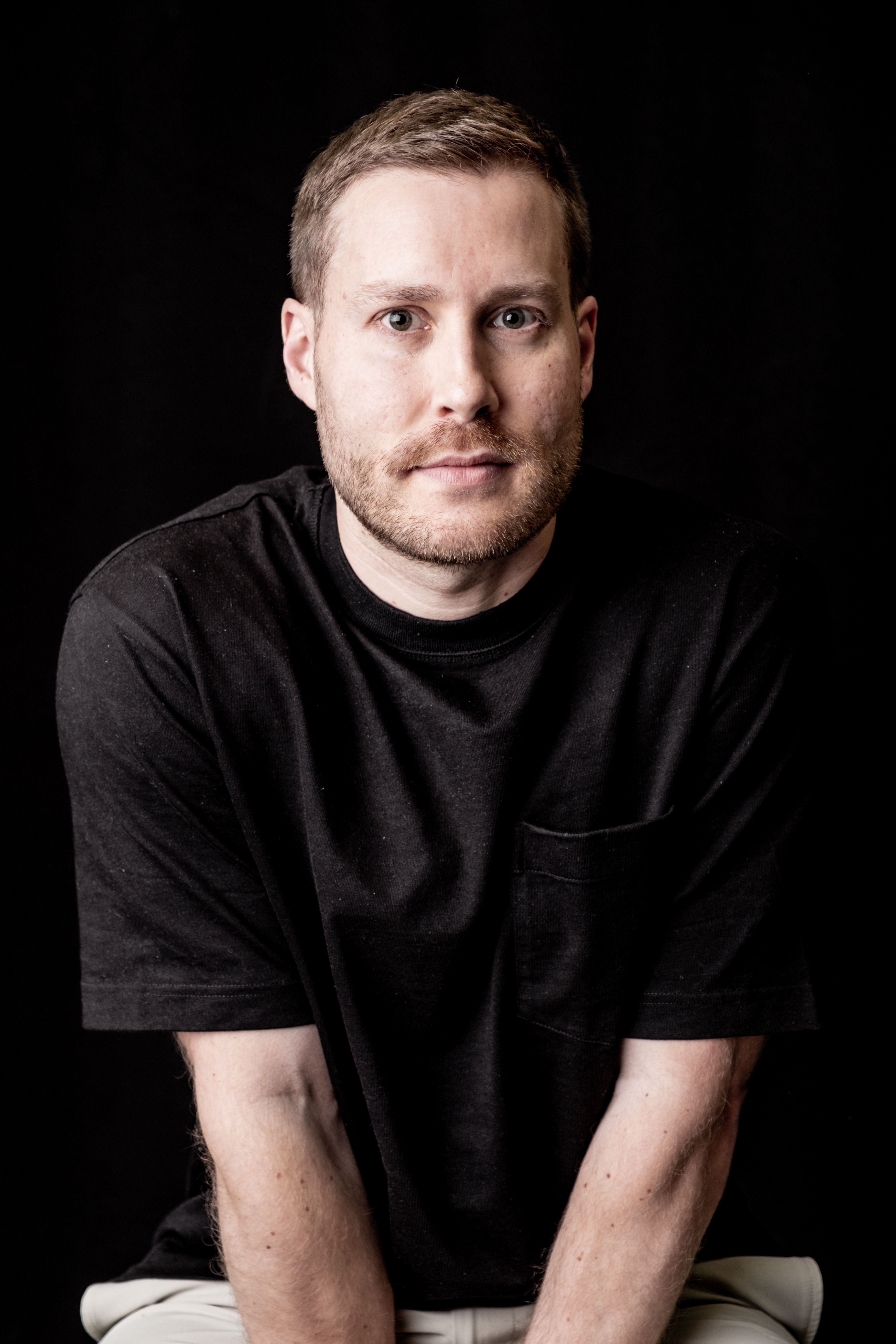A contemplative adult man in a black shirt with a dark background, featuring neutral tones.