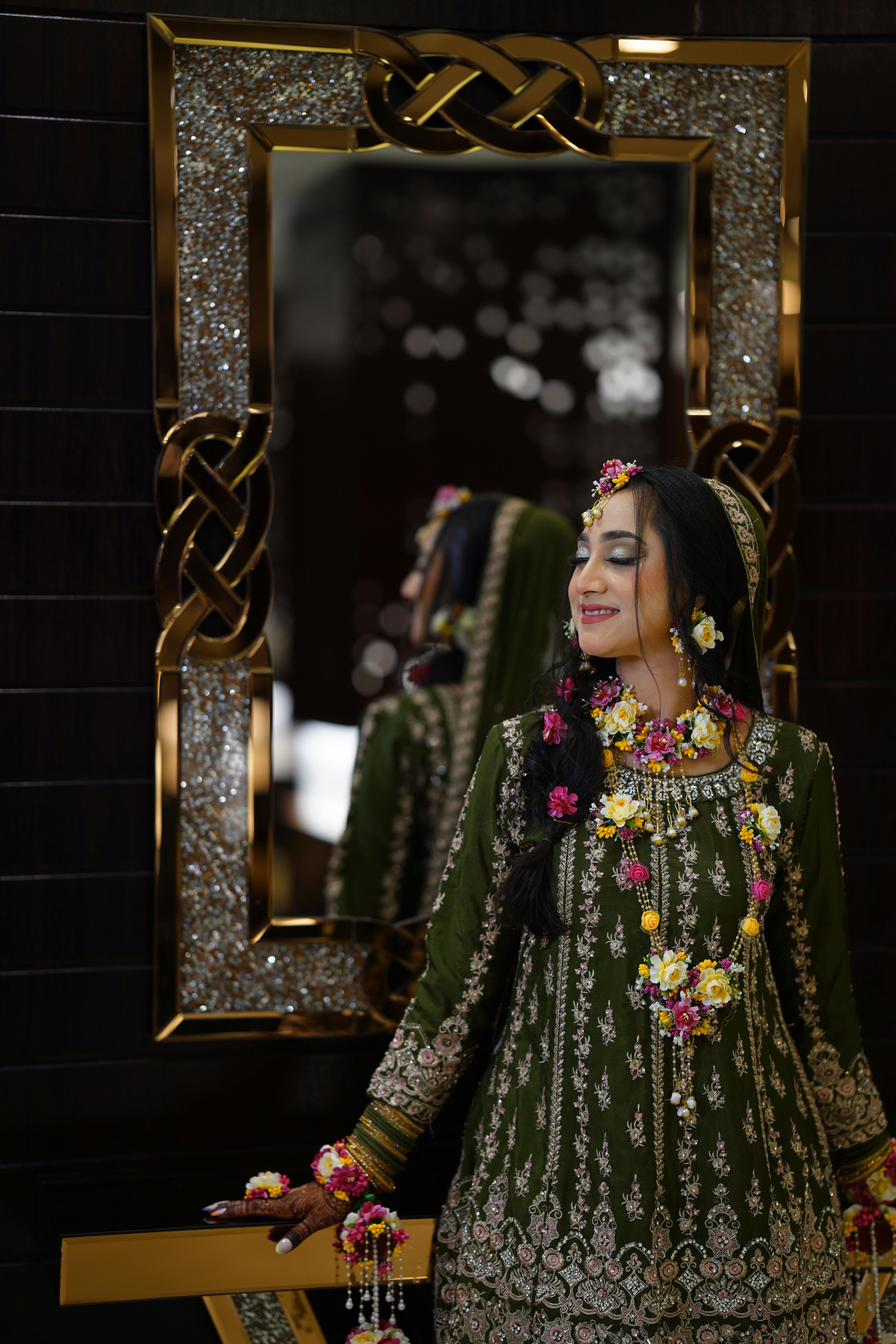 Stunning South Asian bride in green embroidered dress with floral jewelry, standing by decorative mirror.