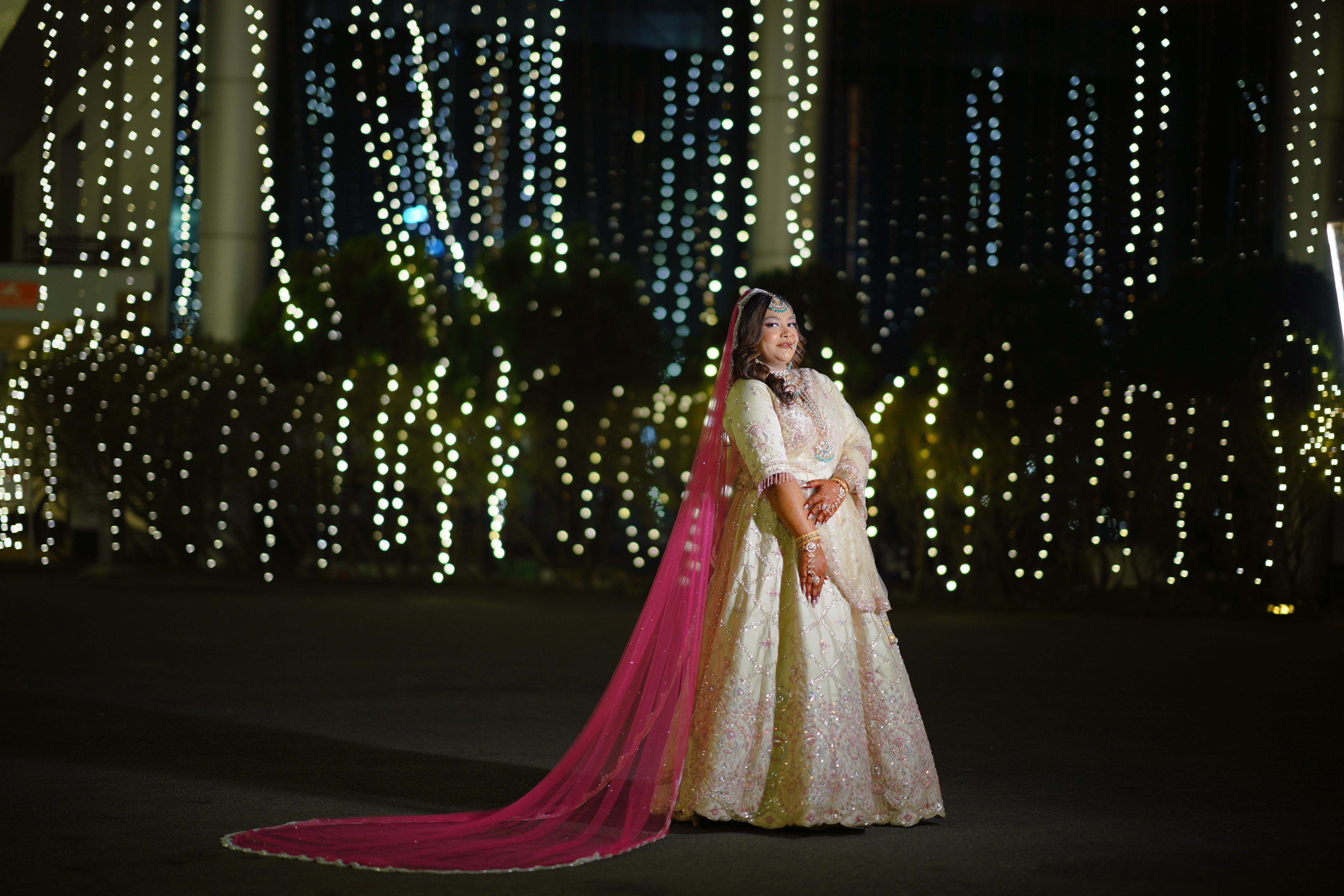 A bride poses elegantly in a sparkling gown with pink veil at a vibrant night wedding, illuminated by twinkling lights.