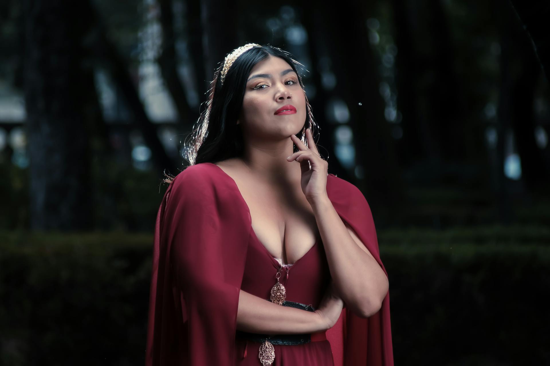 Portrait of a powerful woman in a red dress and gold crown in an outdoor forest setting.