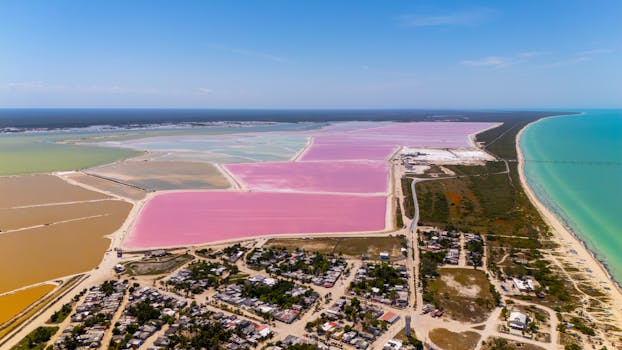 A stunning aerial view of colorful salt flats near a coastal village under a clear blue sky.