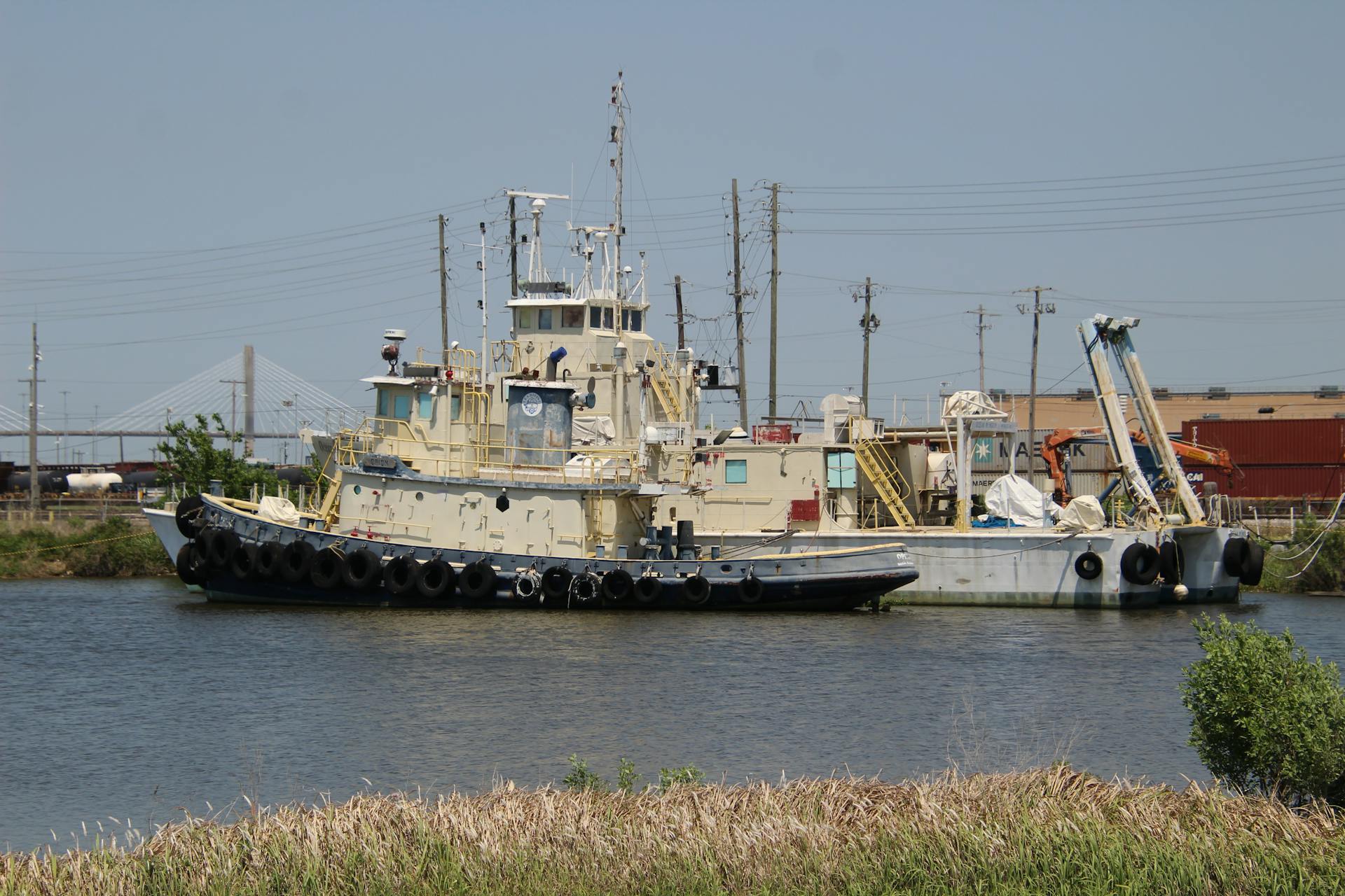 A stationary tugboat and barge beside an industrial harbor under clear blue skies.
