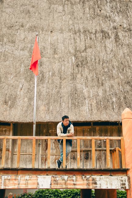A man standing on a traditional balcony in Kon Tum, Vietnam, with a red flag overhead.