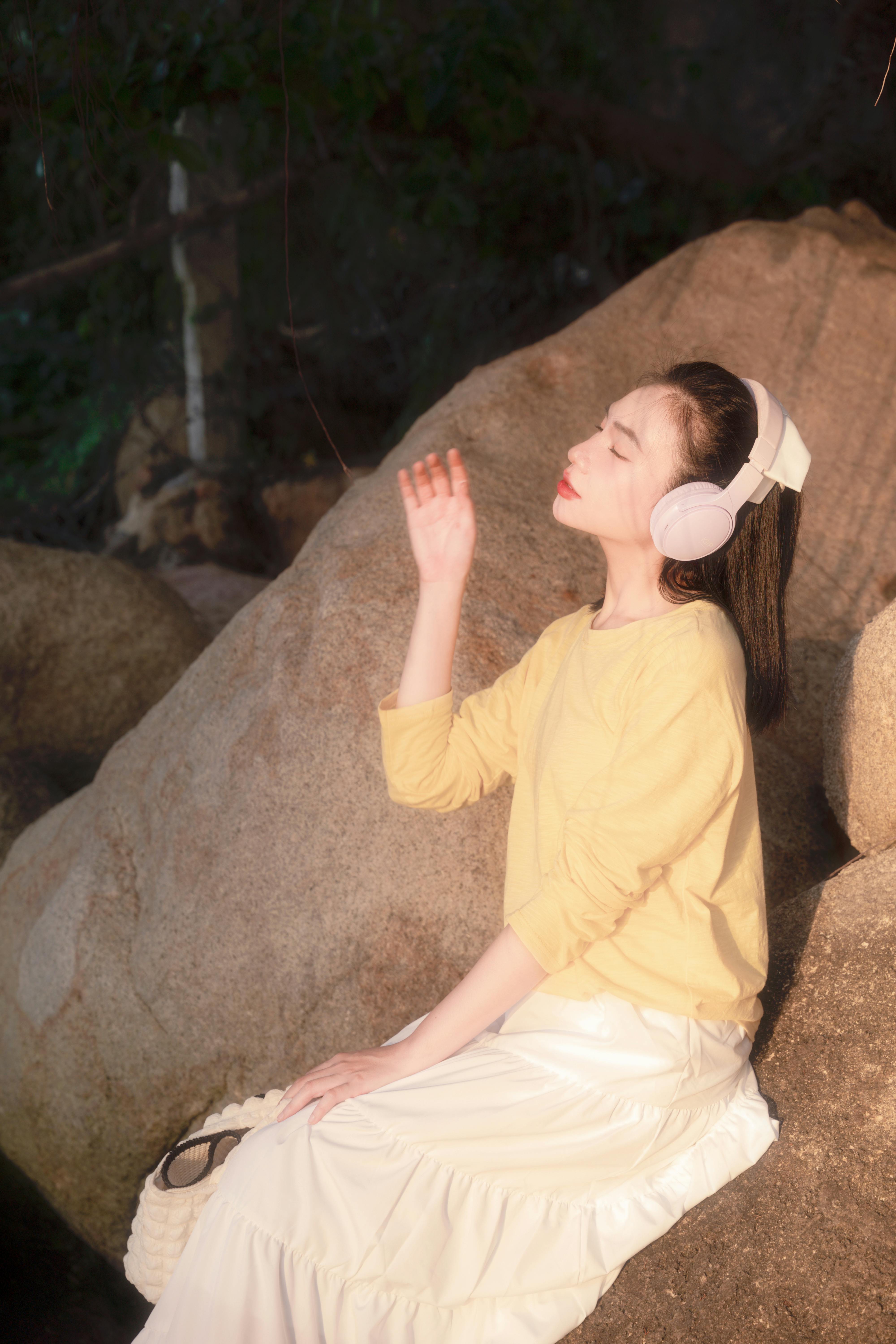 Woman in yellow sweater and white skirt enjoys sunlight outdoors, wearing headphones.