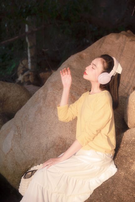 Woman in yellow sweater and white skirt enjoys sunlight outdoors, wearing headphones.