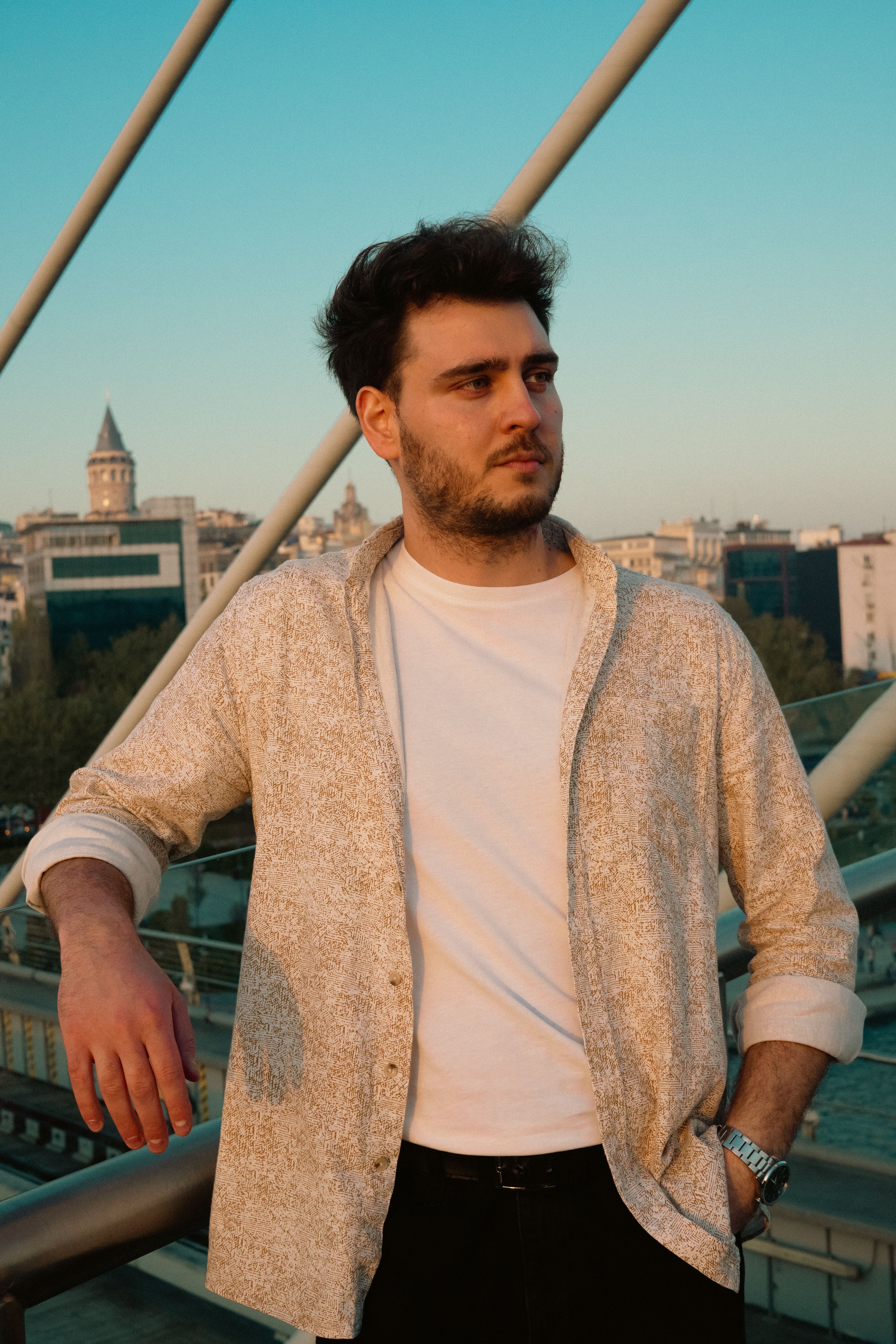 Man in casual attire standing on a bridge with cityscape in the background at sunset.