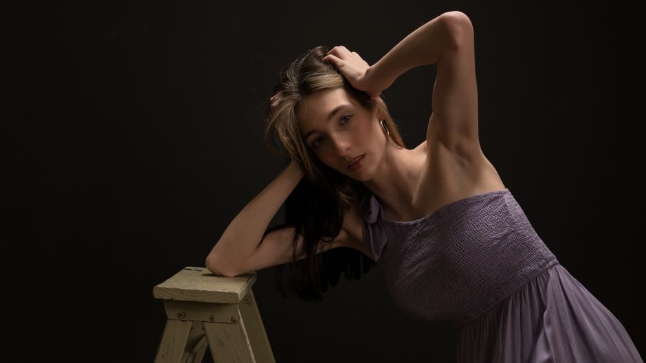 Portrait of a woman in a purple dress posing elegantly with a ladder in a dark studio setting.