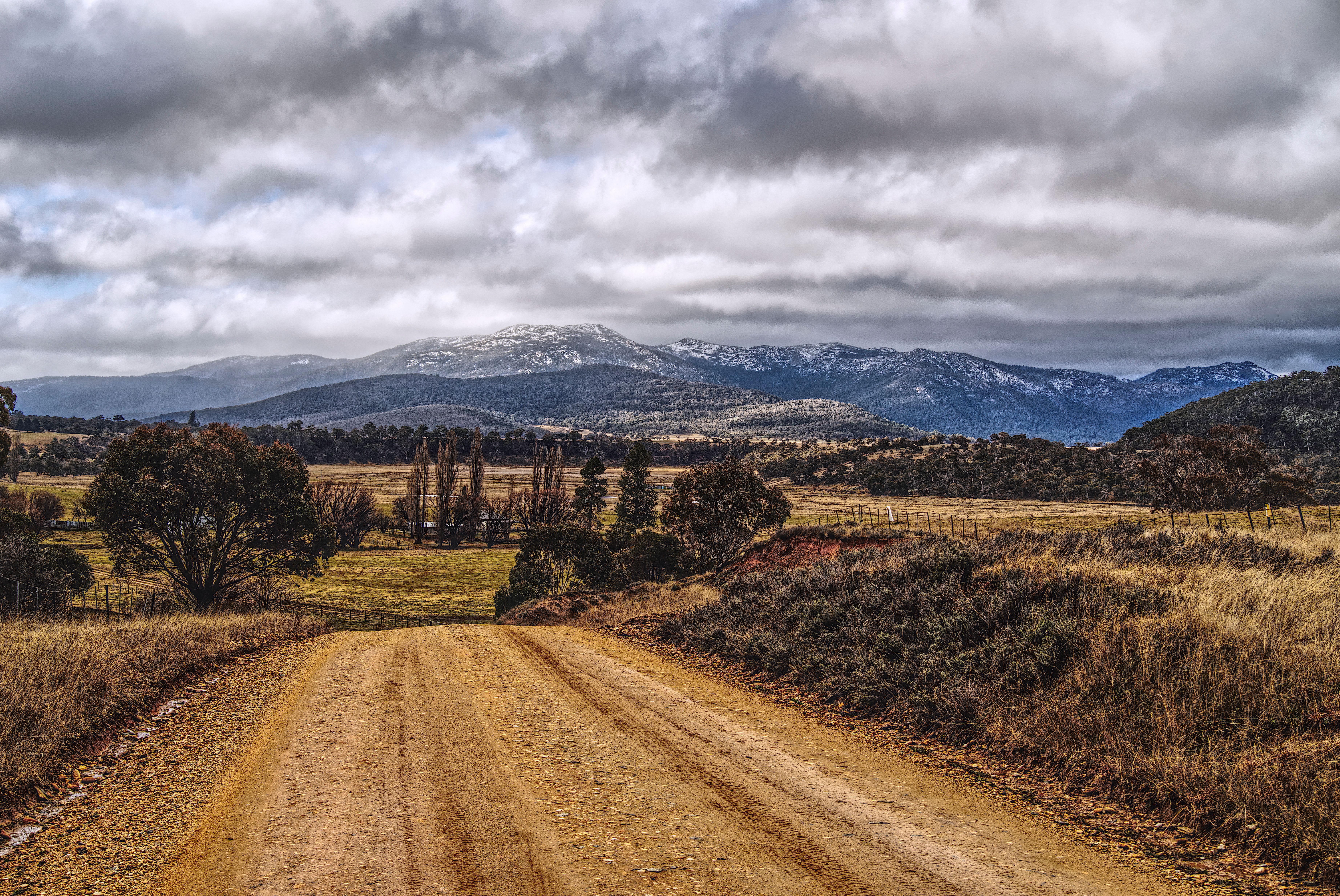 A dirt road leading through countryside landscape to majestic snowcapped mountains under cloudy skies.