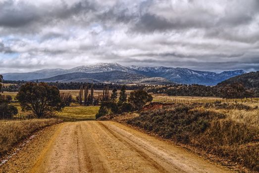 A dirt road leading through countryside landscape to majestic snowcapped mountains under cloudy skies.