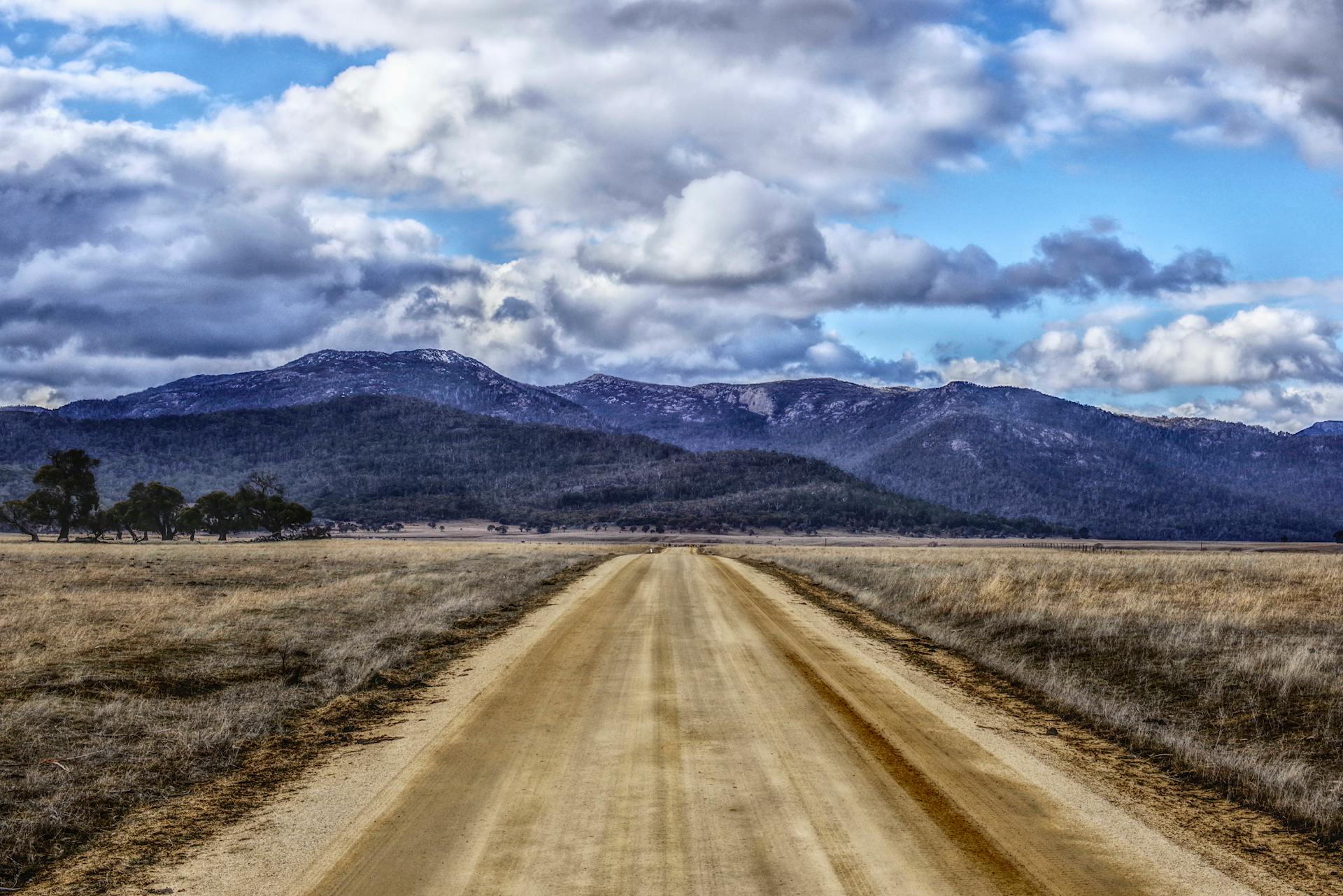 Expansive rural scene with a dirt road heading towards distant mountains under a dramatic sky.