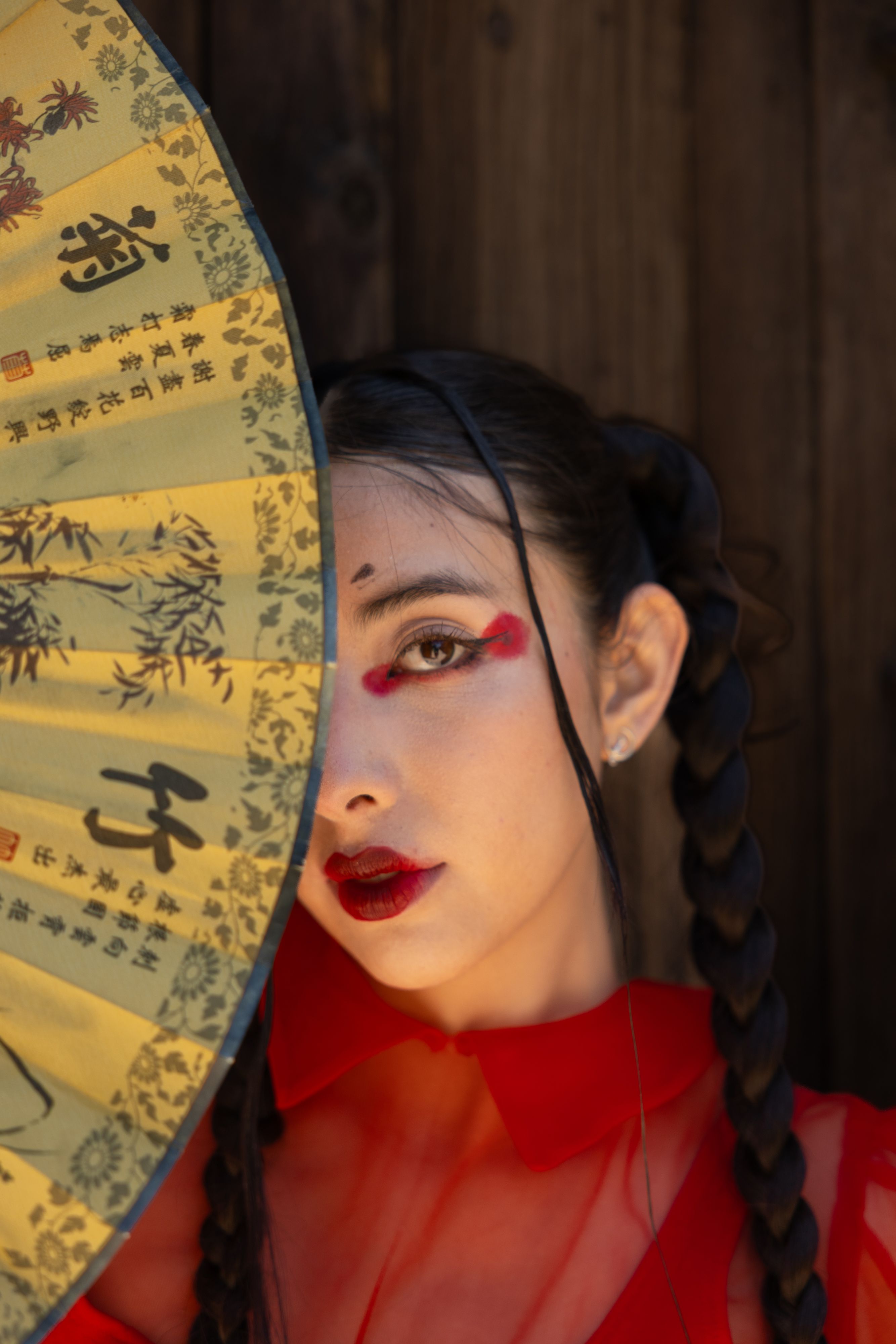 A woman with bold makeup partially hidden by a traditional Asian fan.