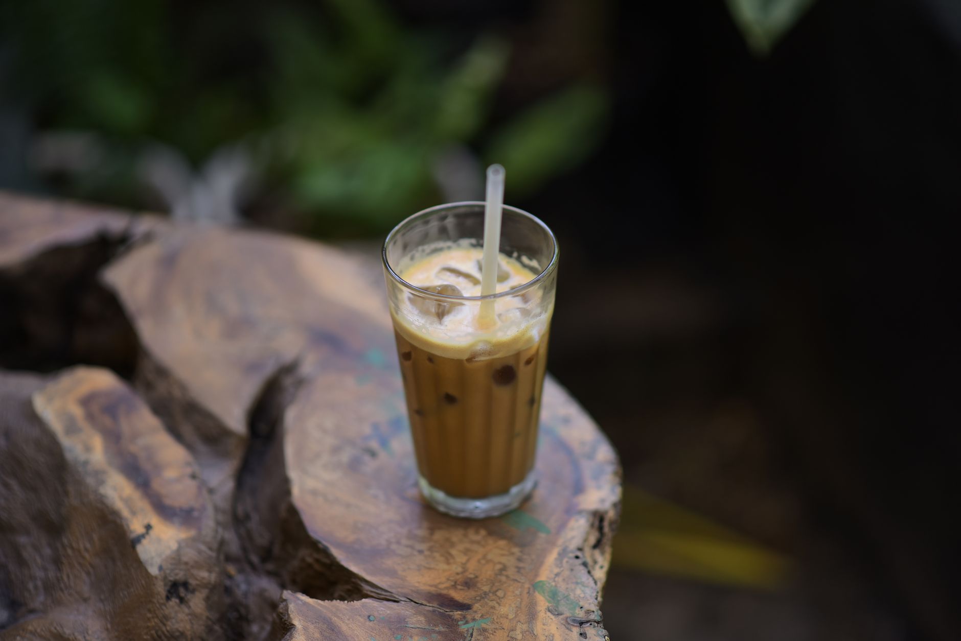 Glass of iced coffee with straw on rustic wooden table in an outdoor setting.