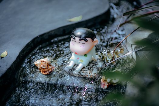 A toy figurine sits peacefully in a flowing water garden, surrounded by shells.
