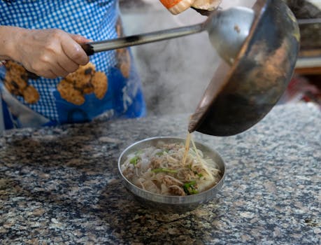 A cook pours steaming broth over noodles in Yunnan, China, capturing local culinary tradition.