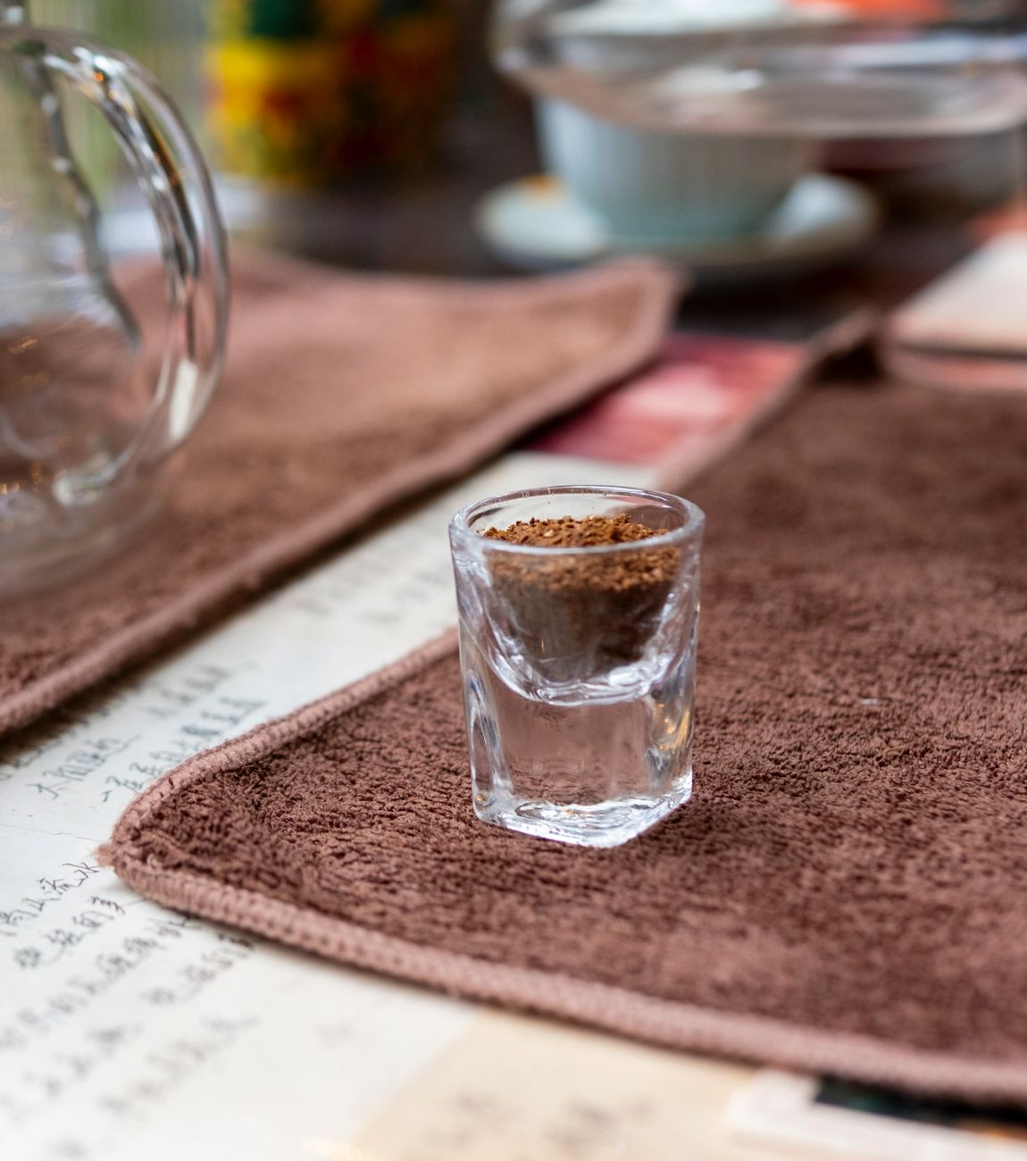 Glass shot filled with coffee grounds on a brown mat, creating a cozy coffee shop vibe.
