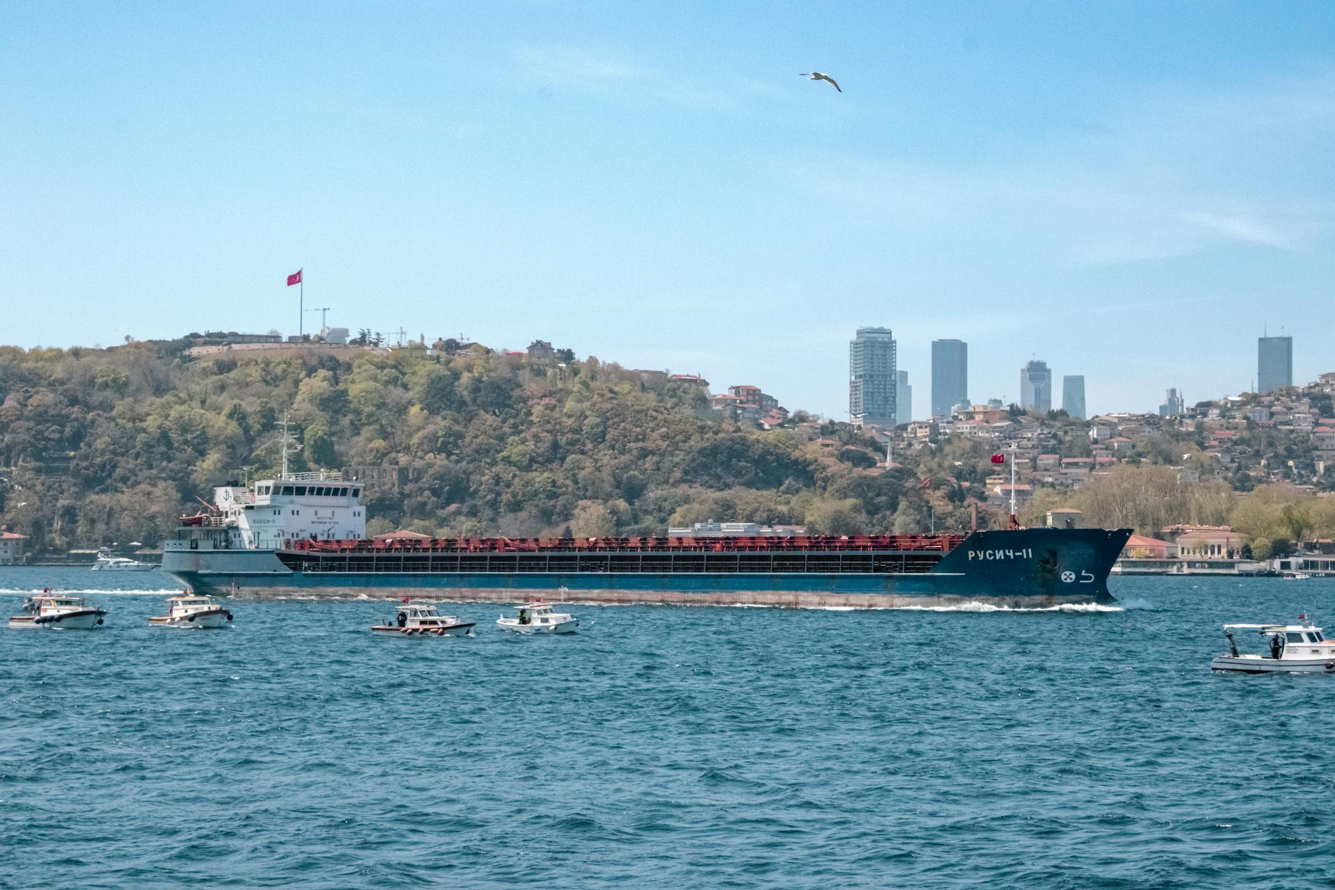 A cargo ship sails through the Bosphorus in Istanbul with a city skyline backdrop.