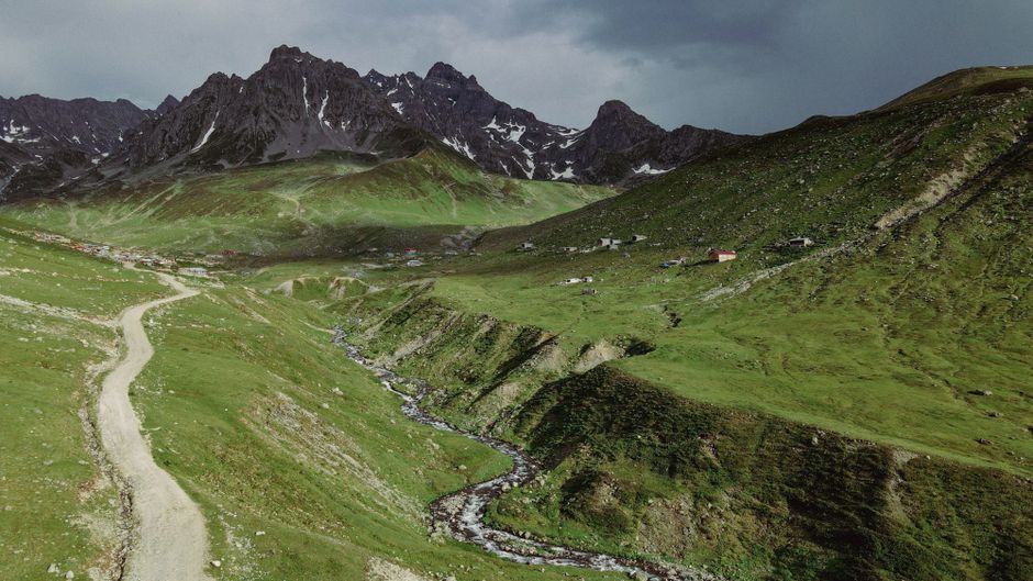 A breathtaking mountain view with a winding stream and dirt trail under a cloudy sky.