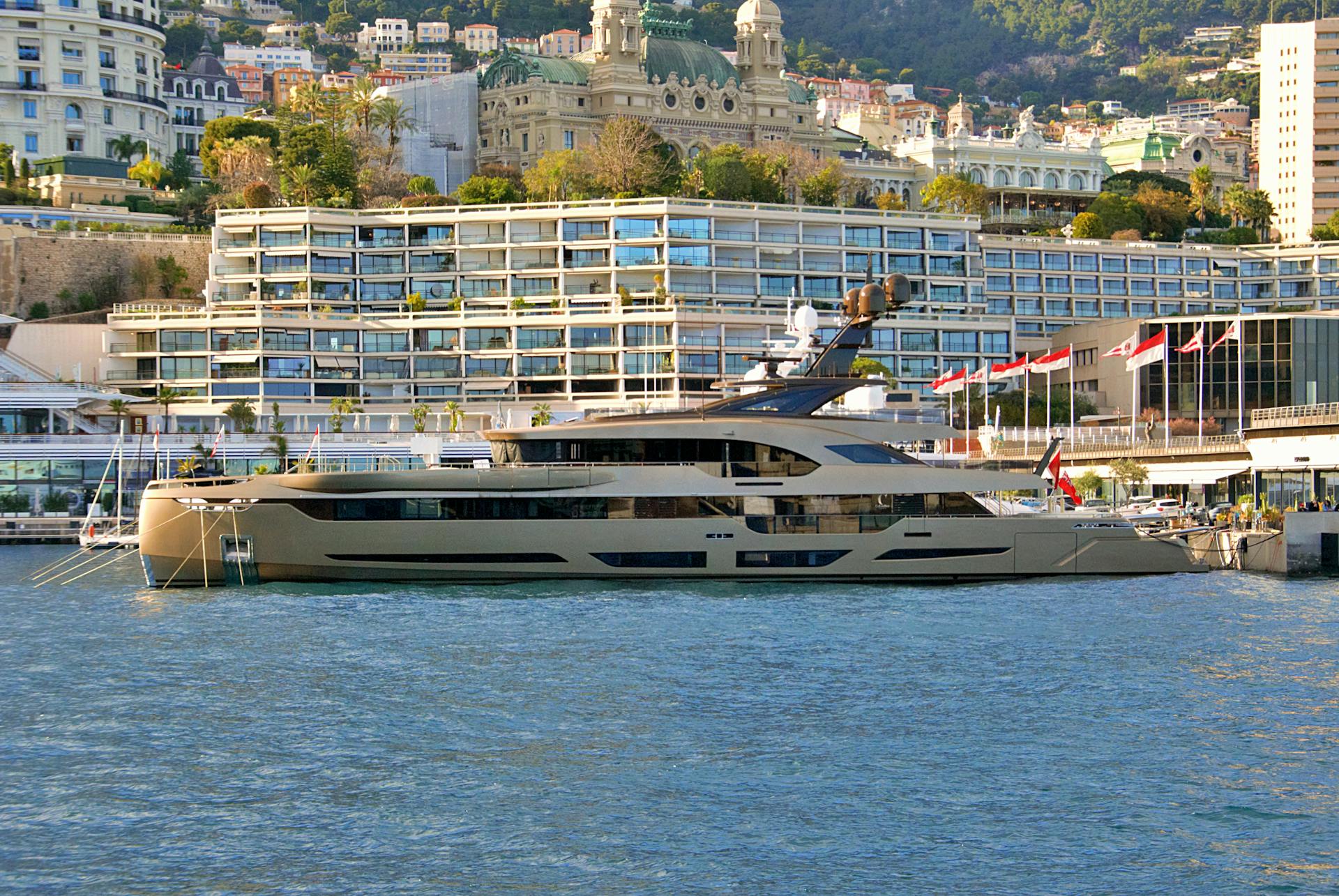 A luxury yacht anchored in Monaco harbor with a stunning view of architecture and flags waving.
