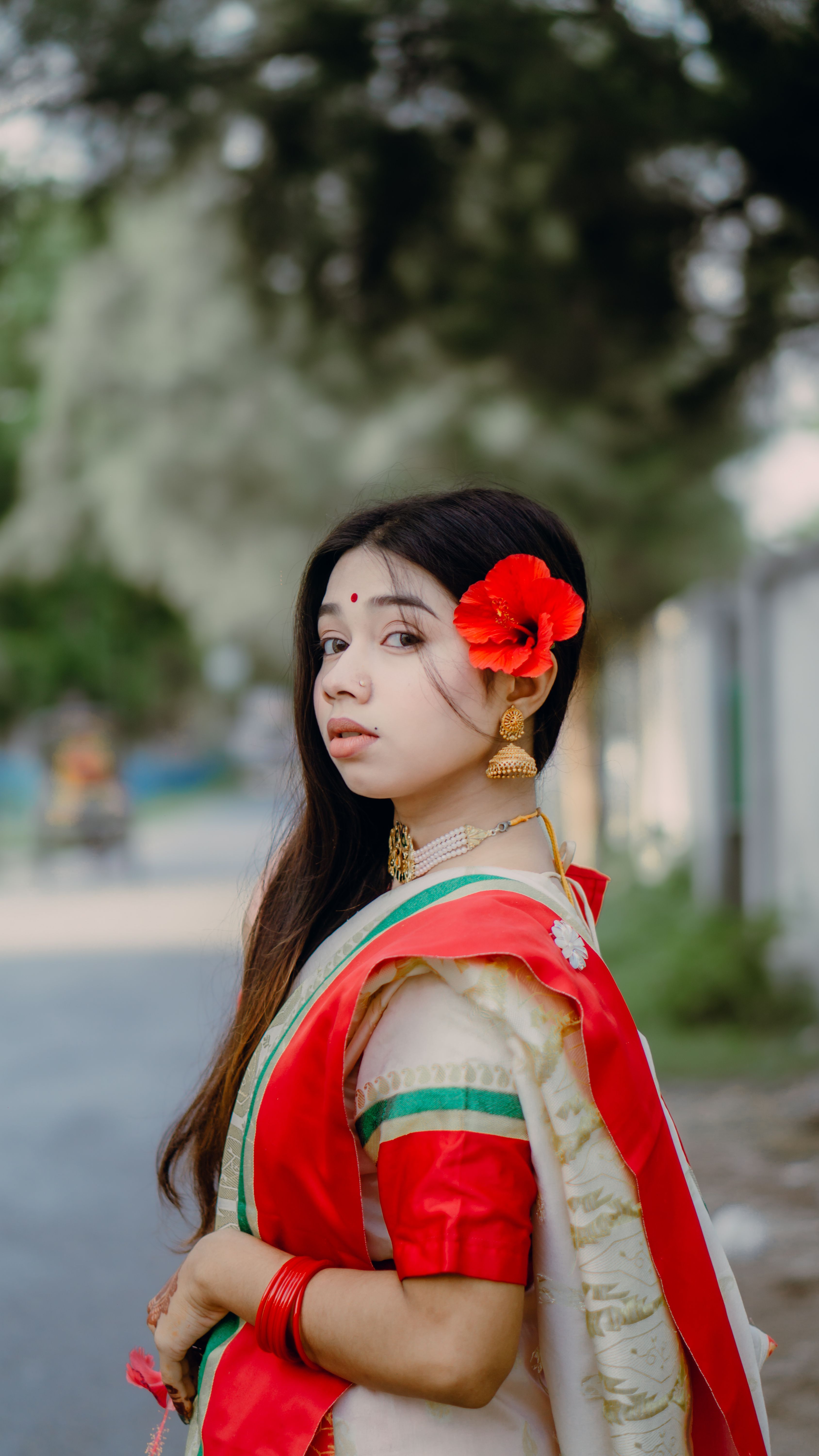 A woman in traditional attire with a red flower in her hair, standing outdoors.
