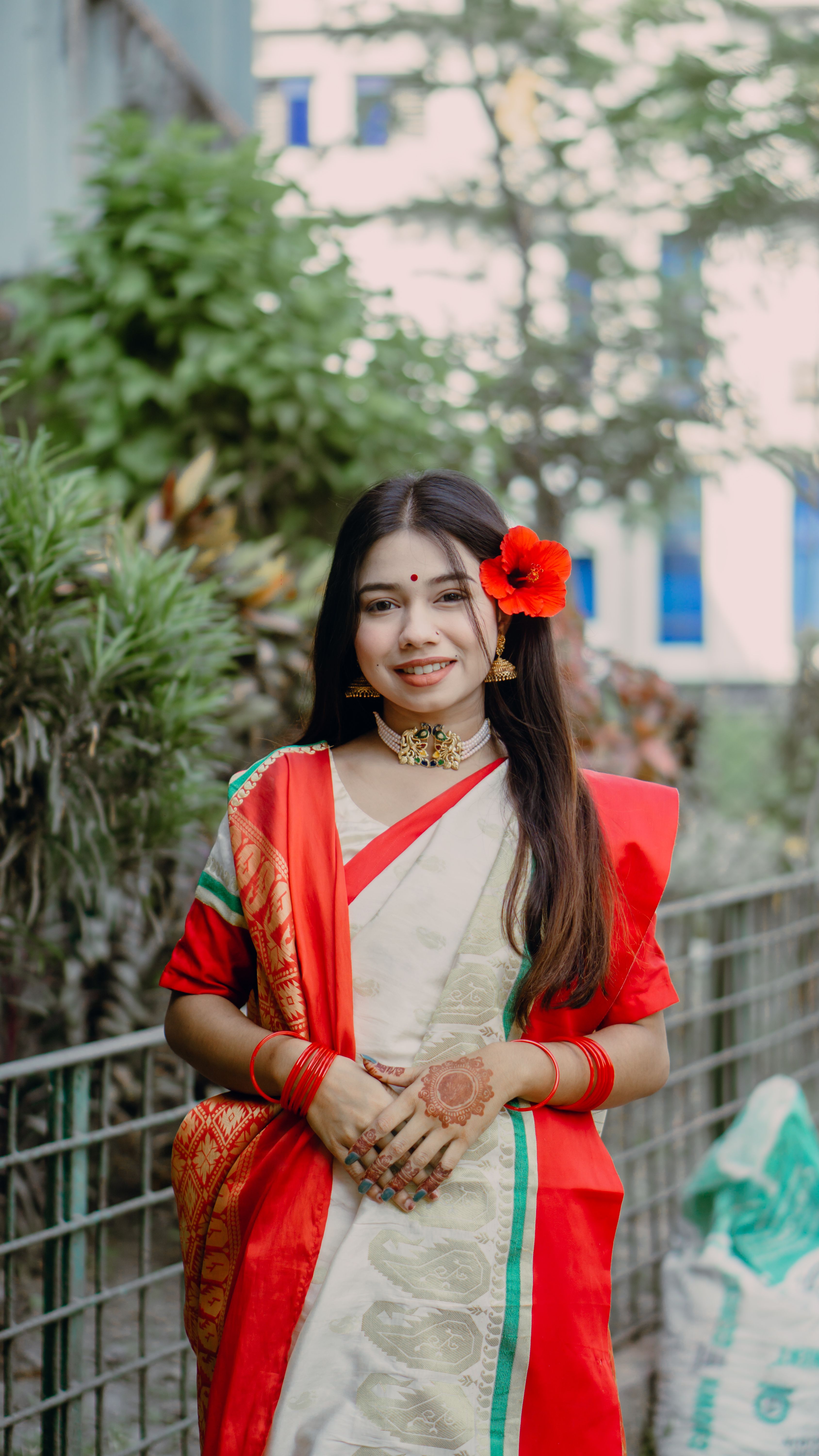 A smiling woman in a red and white sari adorned with jewelry stands outdoors.