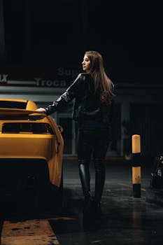A fashionable woman in leather attire leans on a yellow sports car in a dimly lit urban setting.