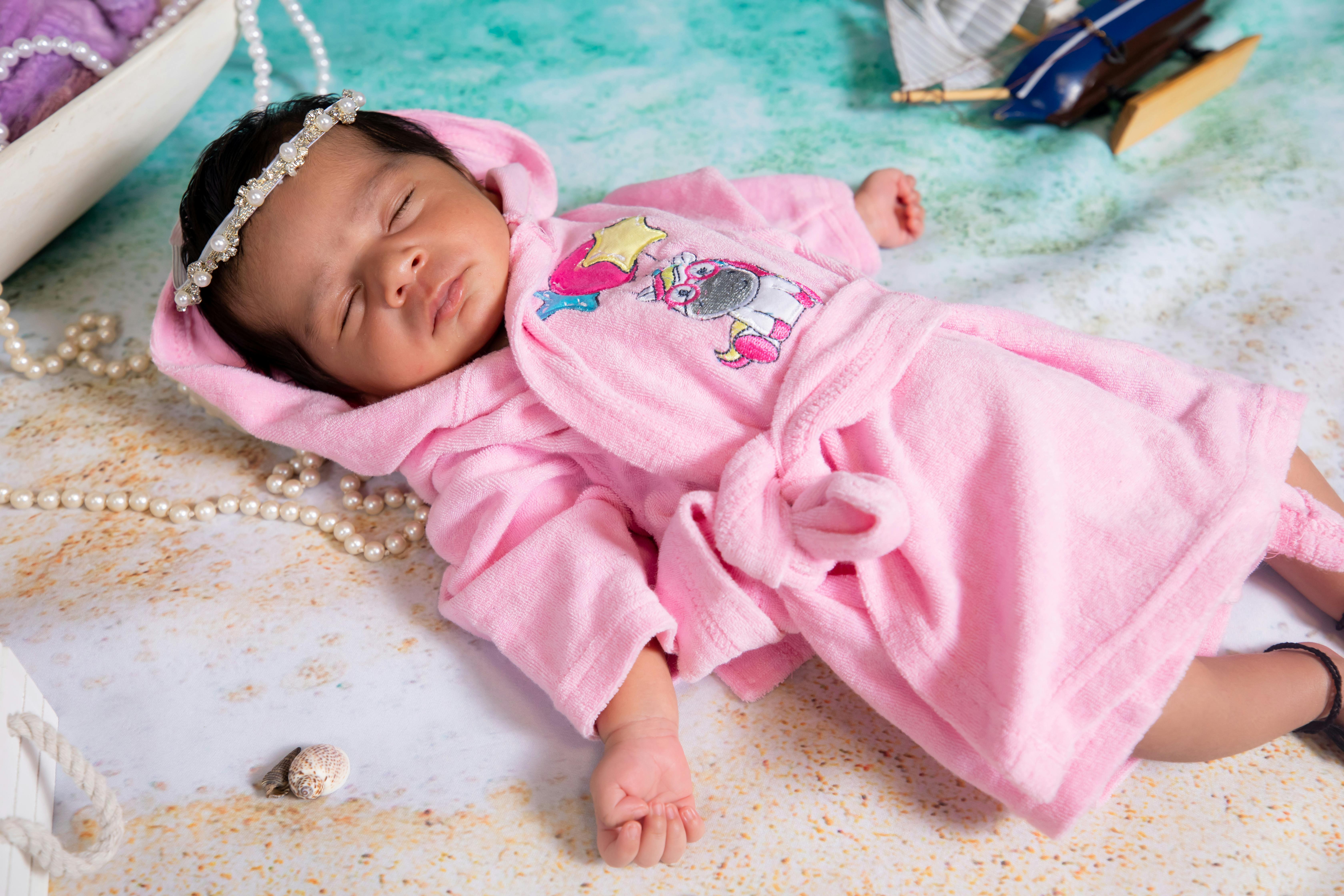 A peaceful newborn Indian baby sleeps in a cute pink robe, surrounded by decorative elements.