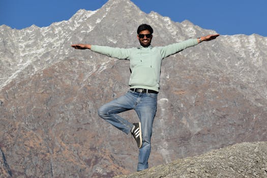 Man in casual attire balancing on one leg against a mountainous backdrop.