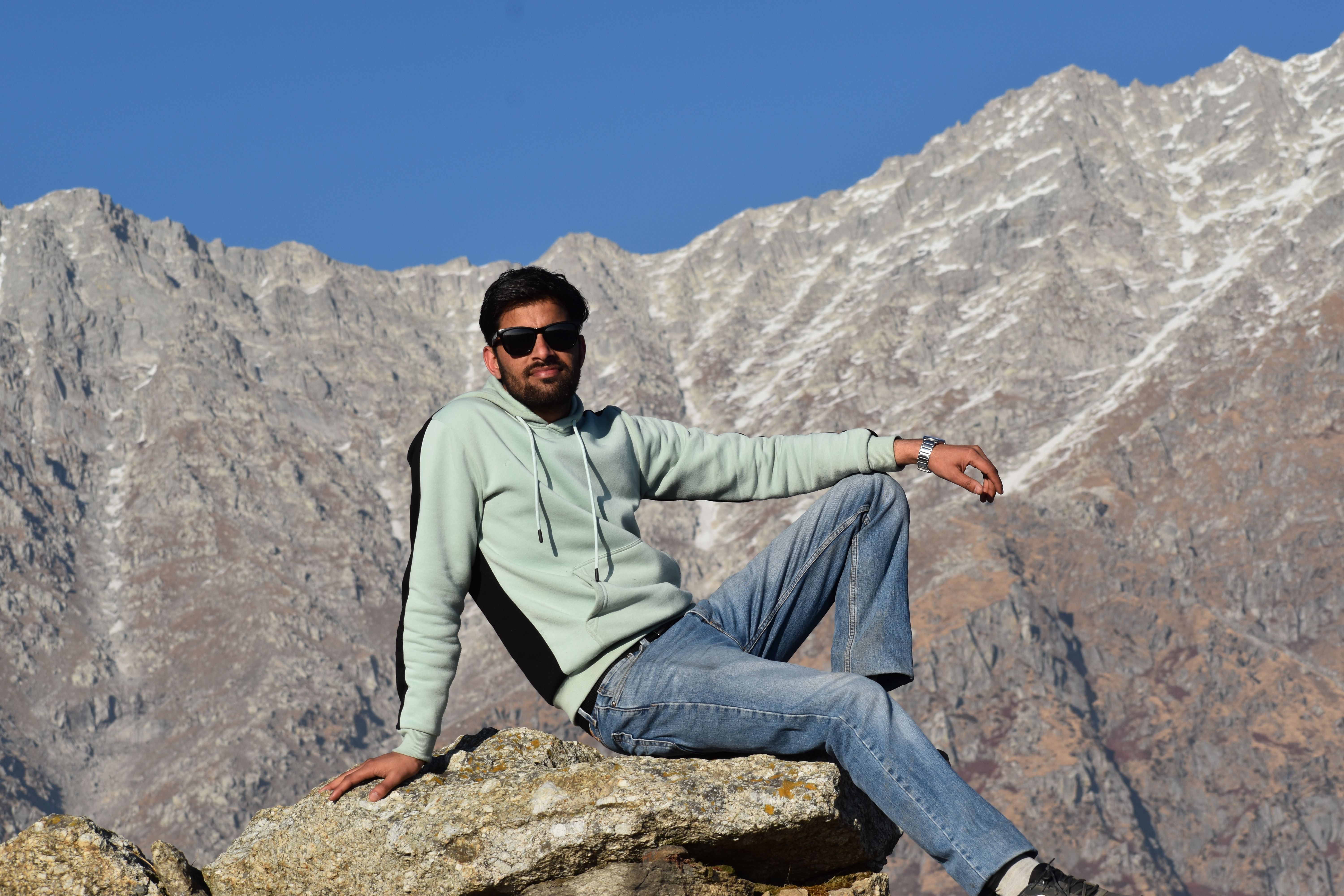 Man sitting on rocky surface against stunning mountain backdrop under clear blue sky