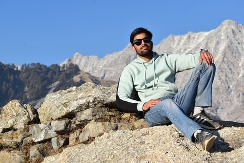 Free Man in sunglasses and casual wear sitting on rocky mountain terrain with a scenic backdrop. Stock Photo