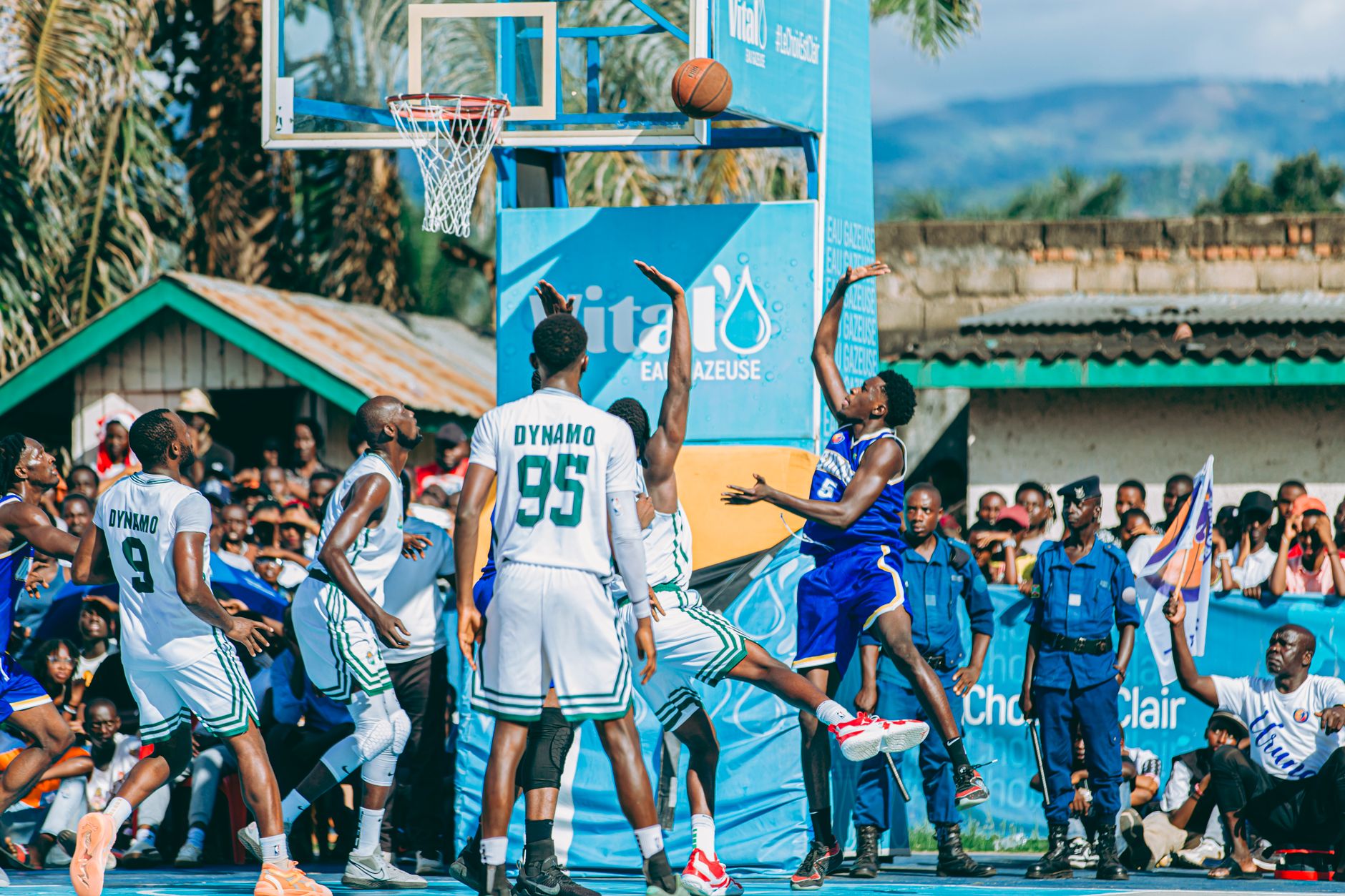 Dynamic outdoor basketball game with players taking a shot and spectators in the background.