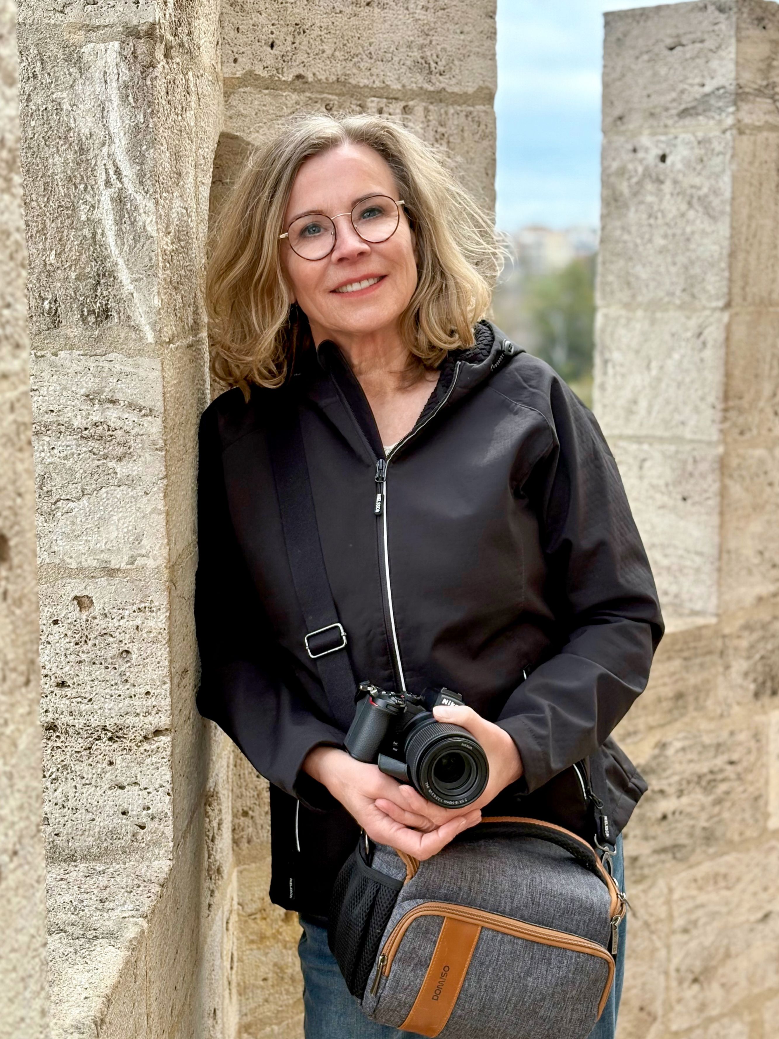 Smiling woman photographer leaning against stone wall, holding camera in Valencia.