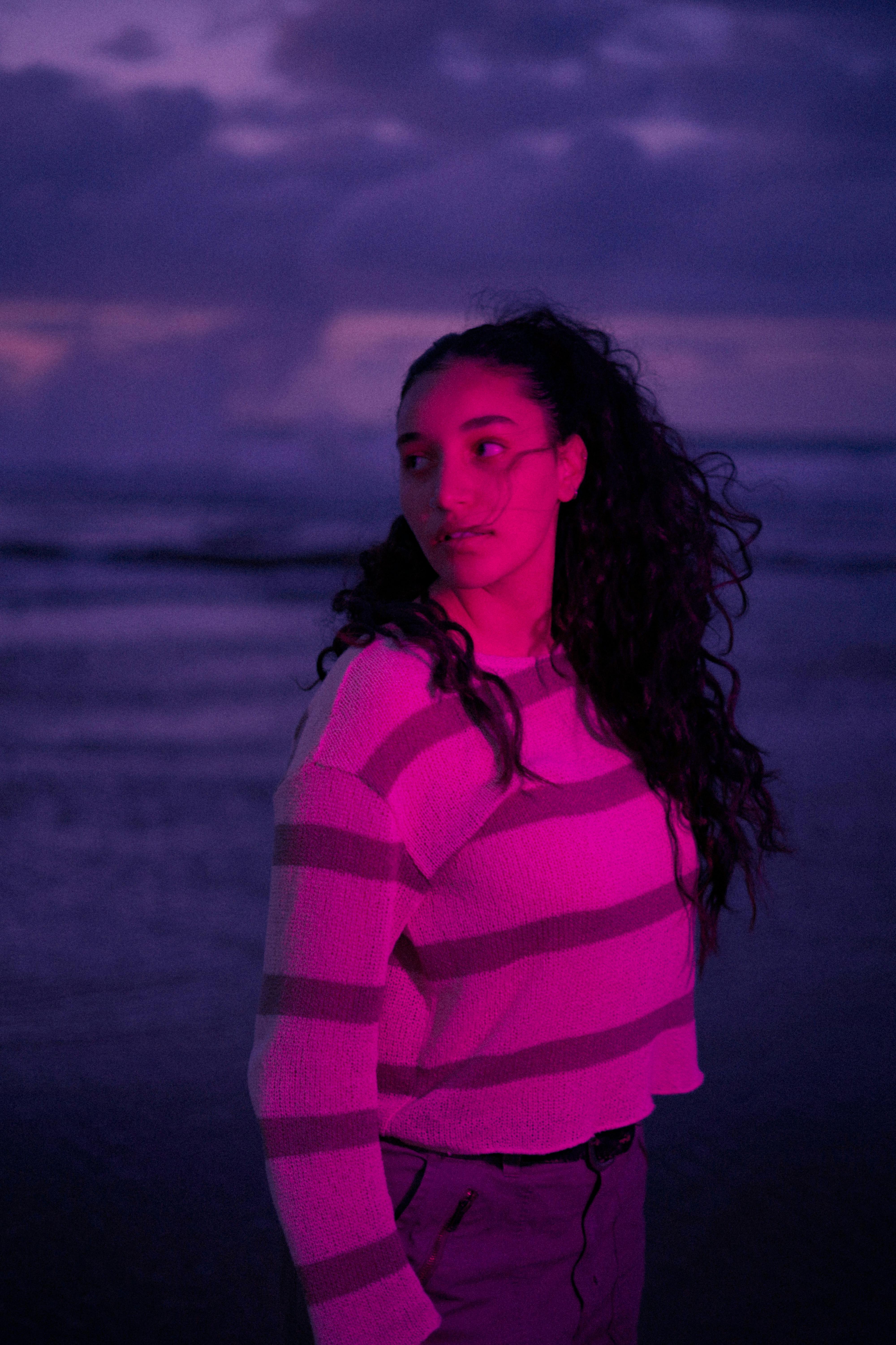 A woman stands on the beach in Rabat under a vibrant twilight sky, casting a pink glow.