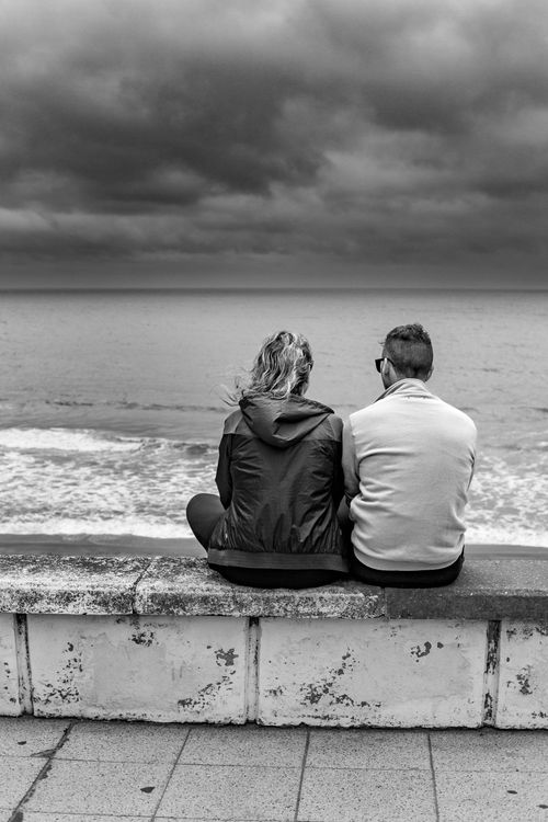 Free A couple sitting on a seawall enjoying the ocean view in Mar del Plata under a dramatic sky. Stock Photo
