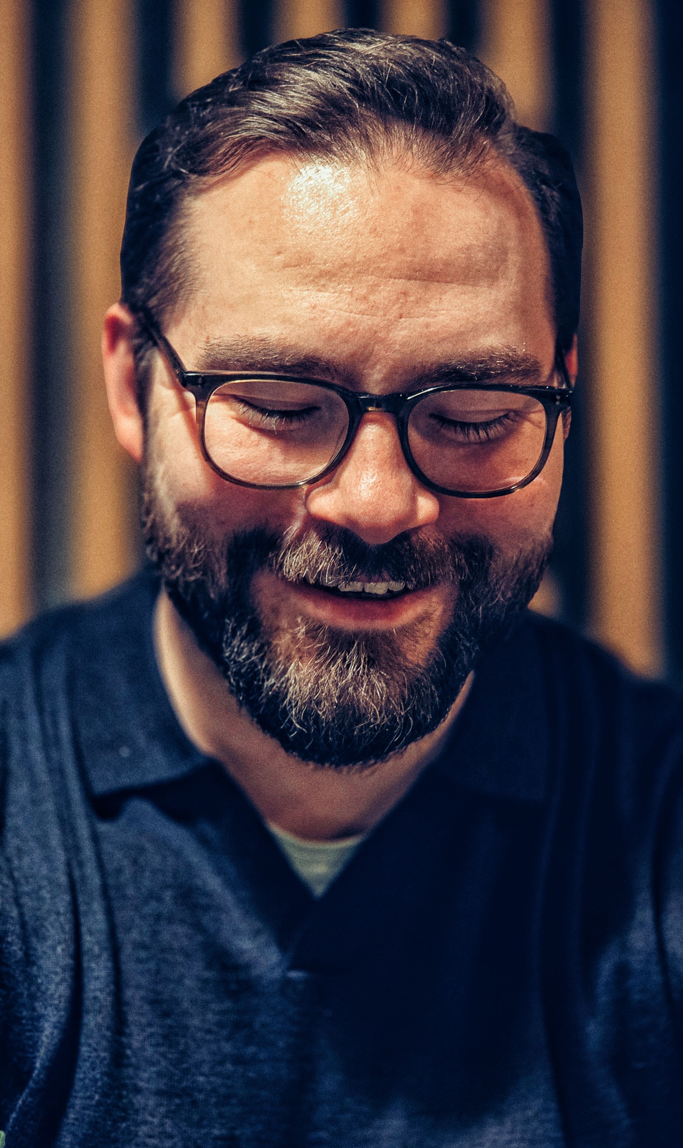 Bearded man with glasses smiles warmly inside a cozy cafe. Warm tones and friendly atmosphere.