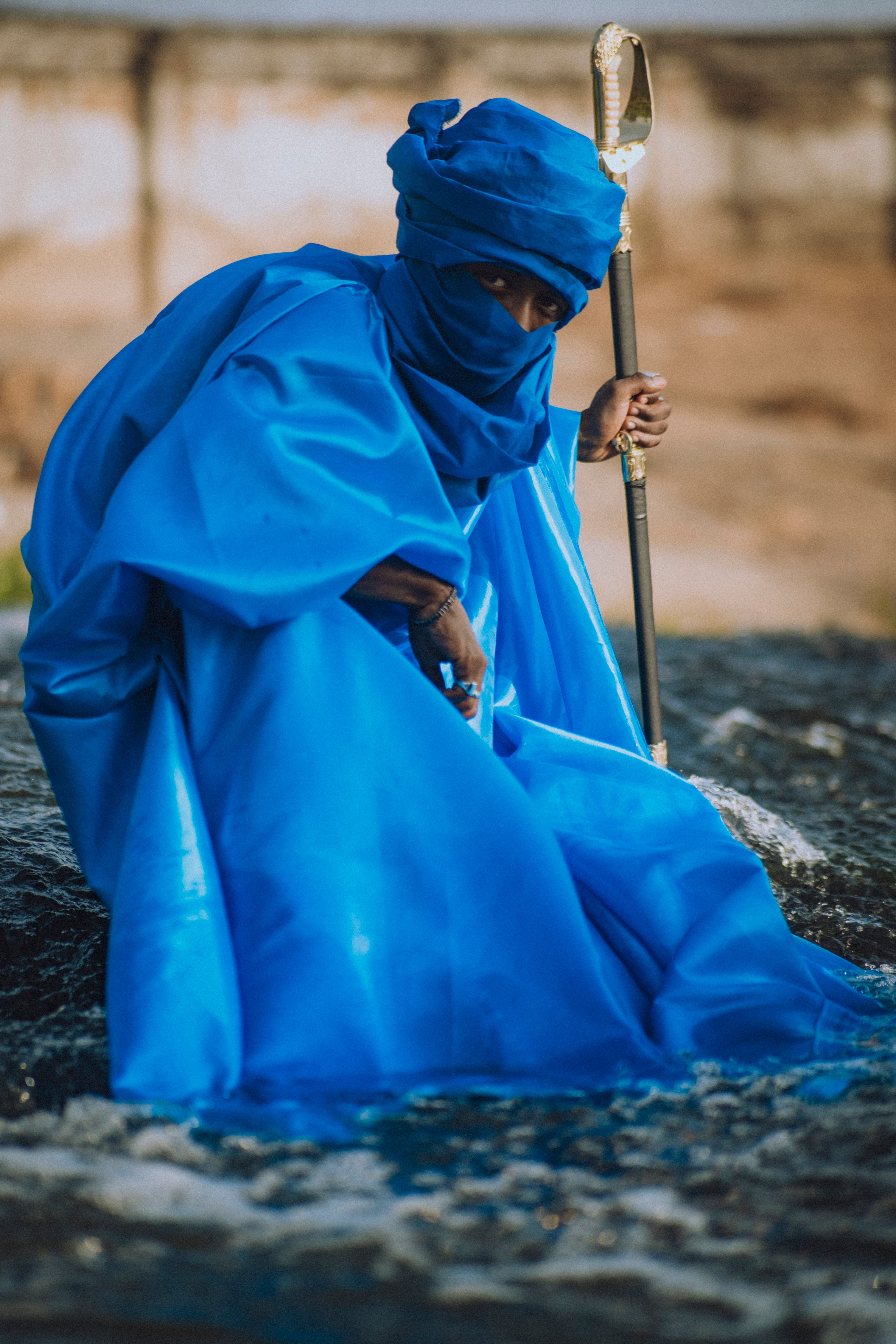 A person in bright blue attire, holding a staff, set against an outdoor landscape.