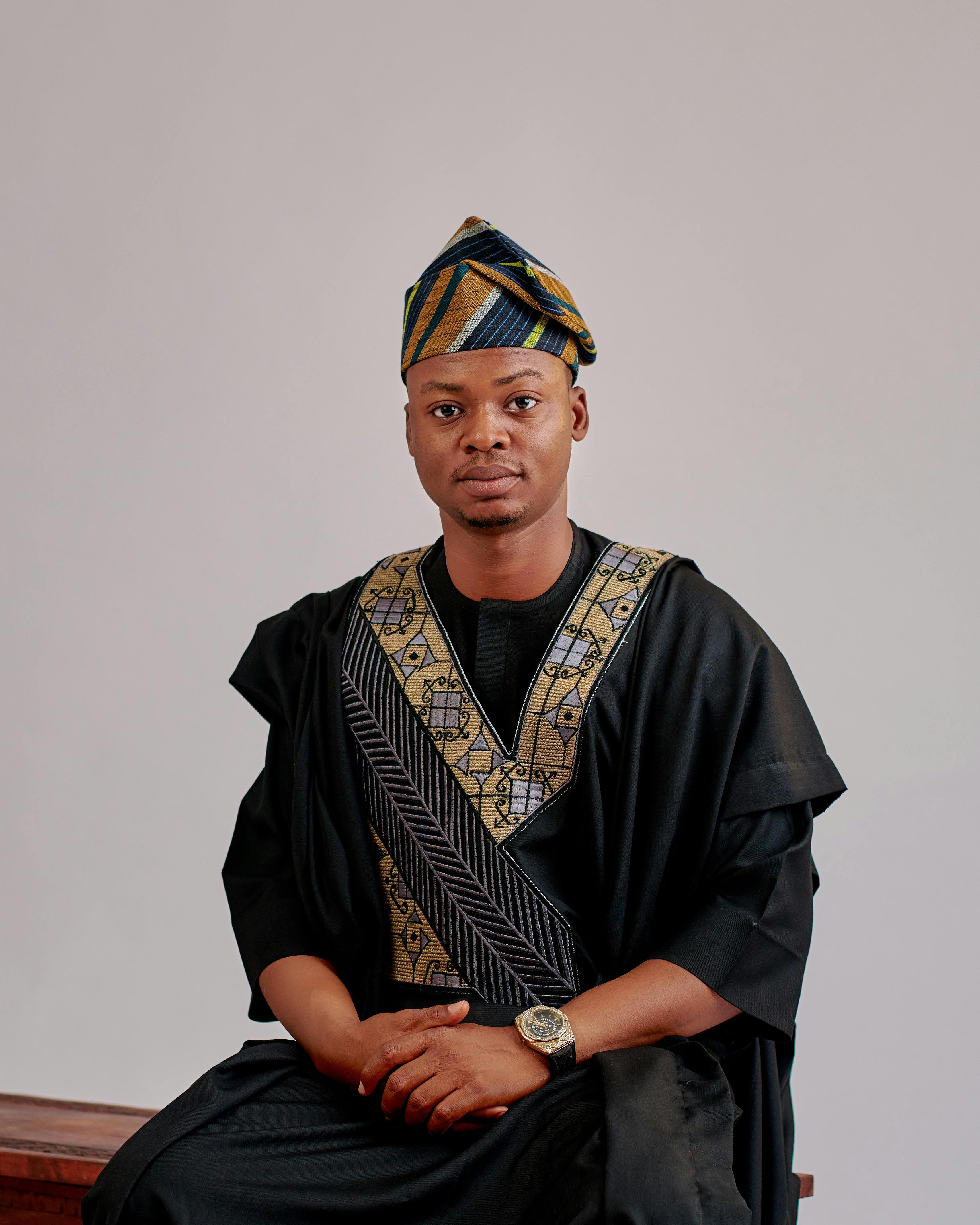 Adult male in Nigerian Agbada sits against a plain background, wearing traditional Yoruba attire.