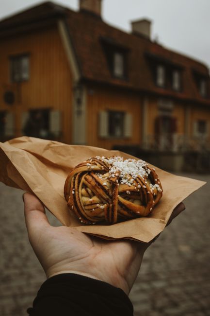 Hand holding a cinnamon roll in a scenic Stockholm street, showcasing Swedish pastry culture.