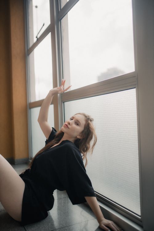 Free A young woman in a black outfit poses gracefully by a large window in soft natural light. Stock Photo