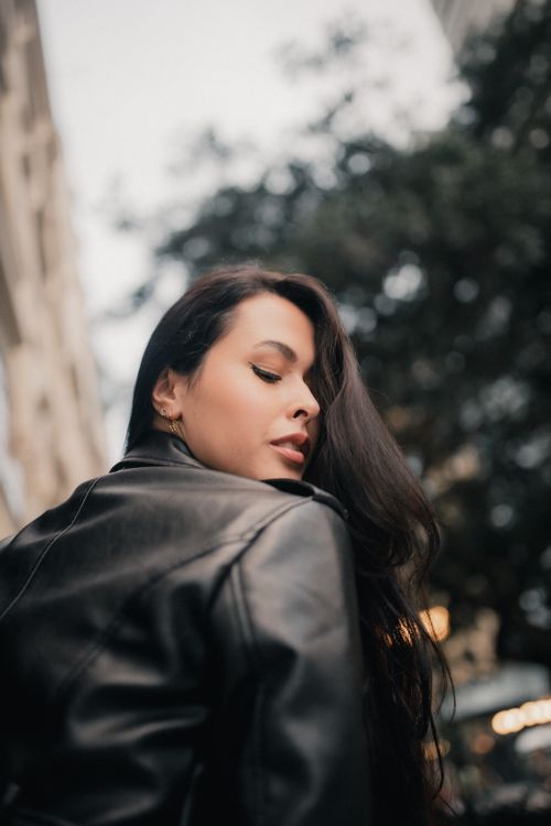 Free Captivating street portrait of a woman in a black leather jacket looking back with cityscape background. Stock Photo