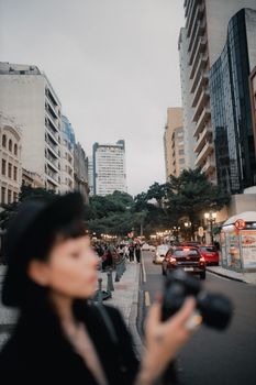 Blurred photographer on bustling city street with tall buildings and trees in the background.