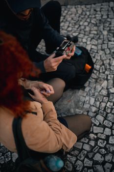 Two adults sitting on a cobblestone street using a recording device, suggesting a musical or journalistic activity.