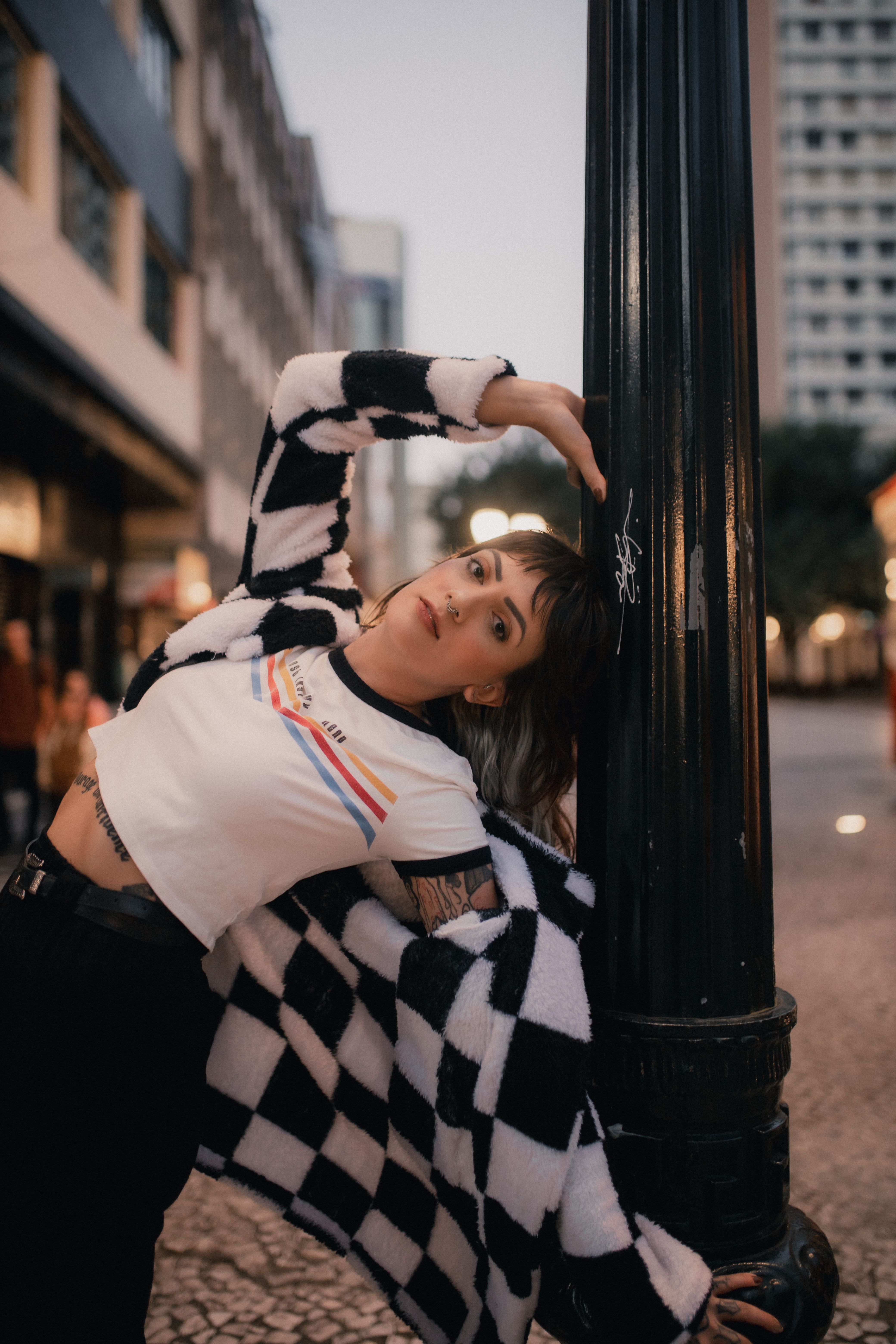 Fashionable woman in checkered coat leans stylishly against a lamppost on an urban street.